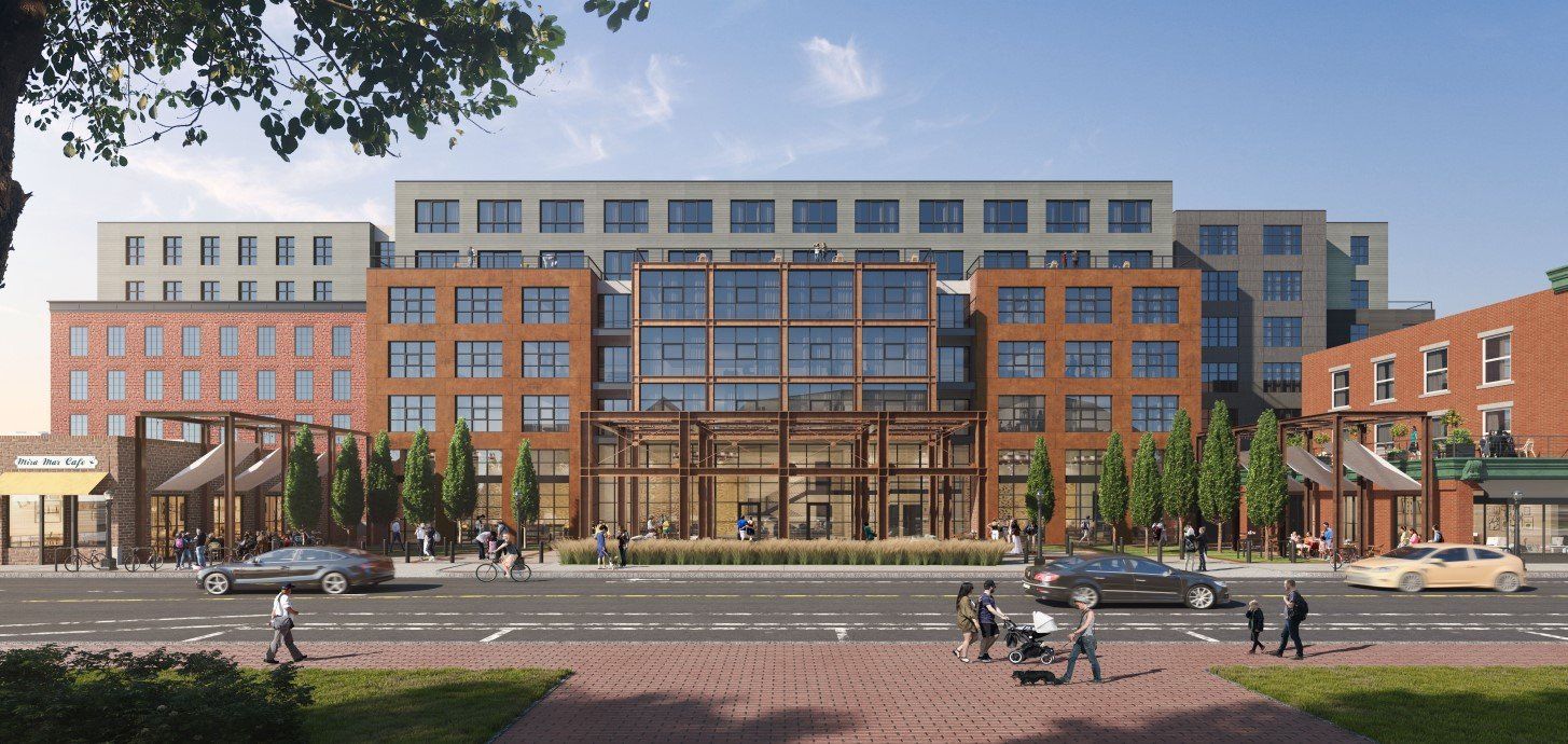 A multi-story brick and grey building featuring a glass-fronted entrance, trees, street traffic, and pedestrians.