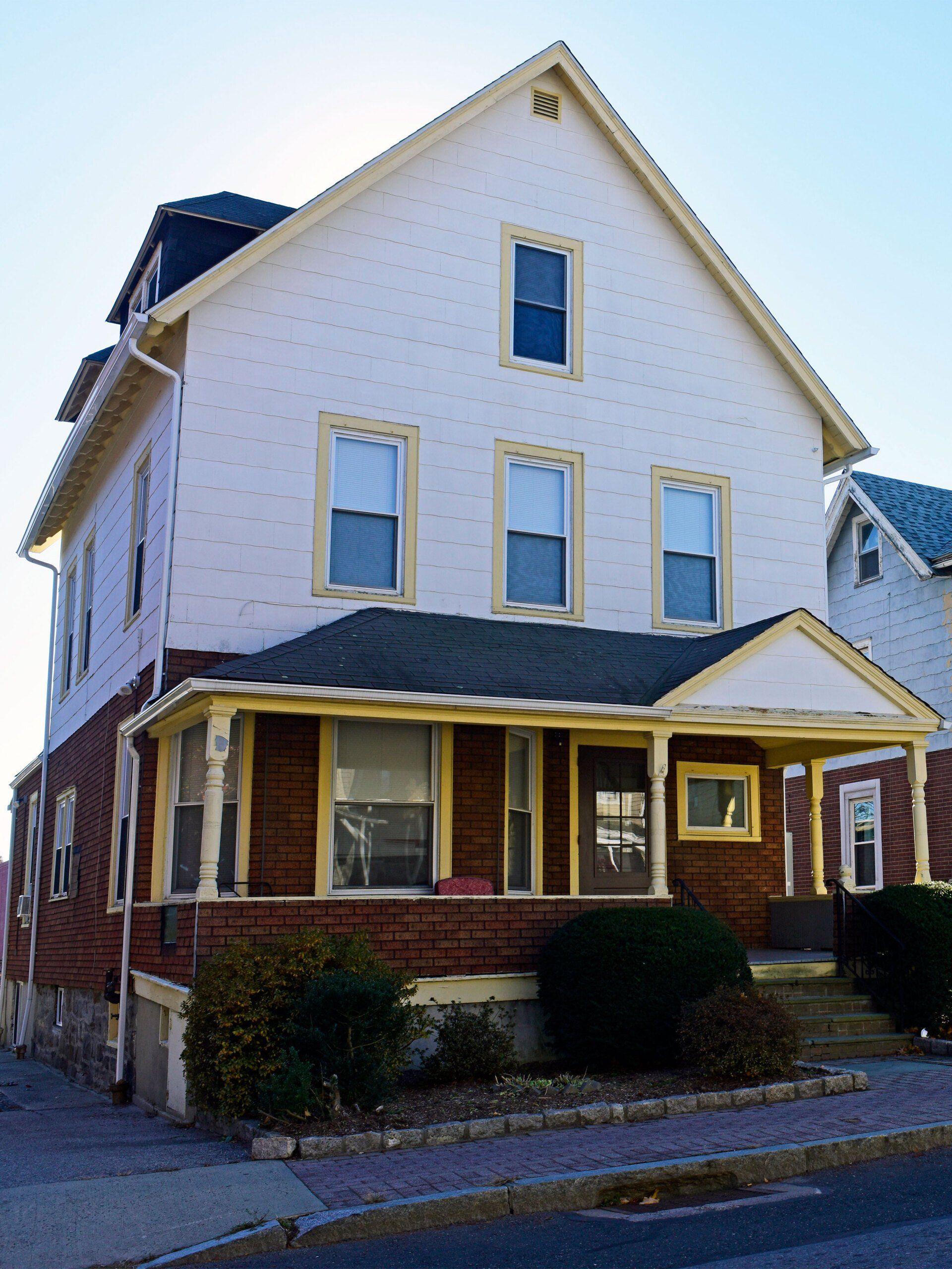 A three-story house with white siding and a brick-faced lower level, featuring yellow trim and a front porch.