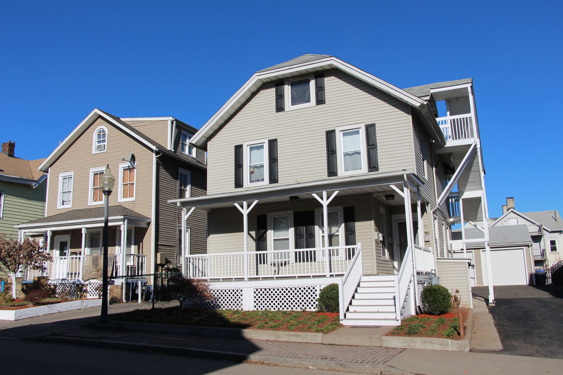 Two tan houses with front porches and white trim stand side-by-side under a clear blue sky.