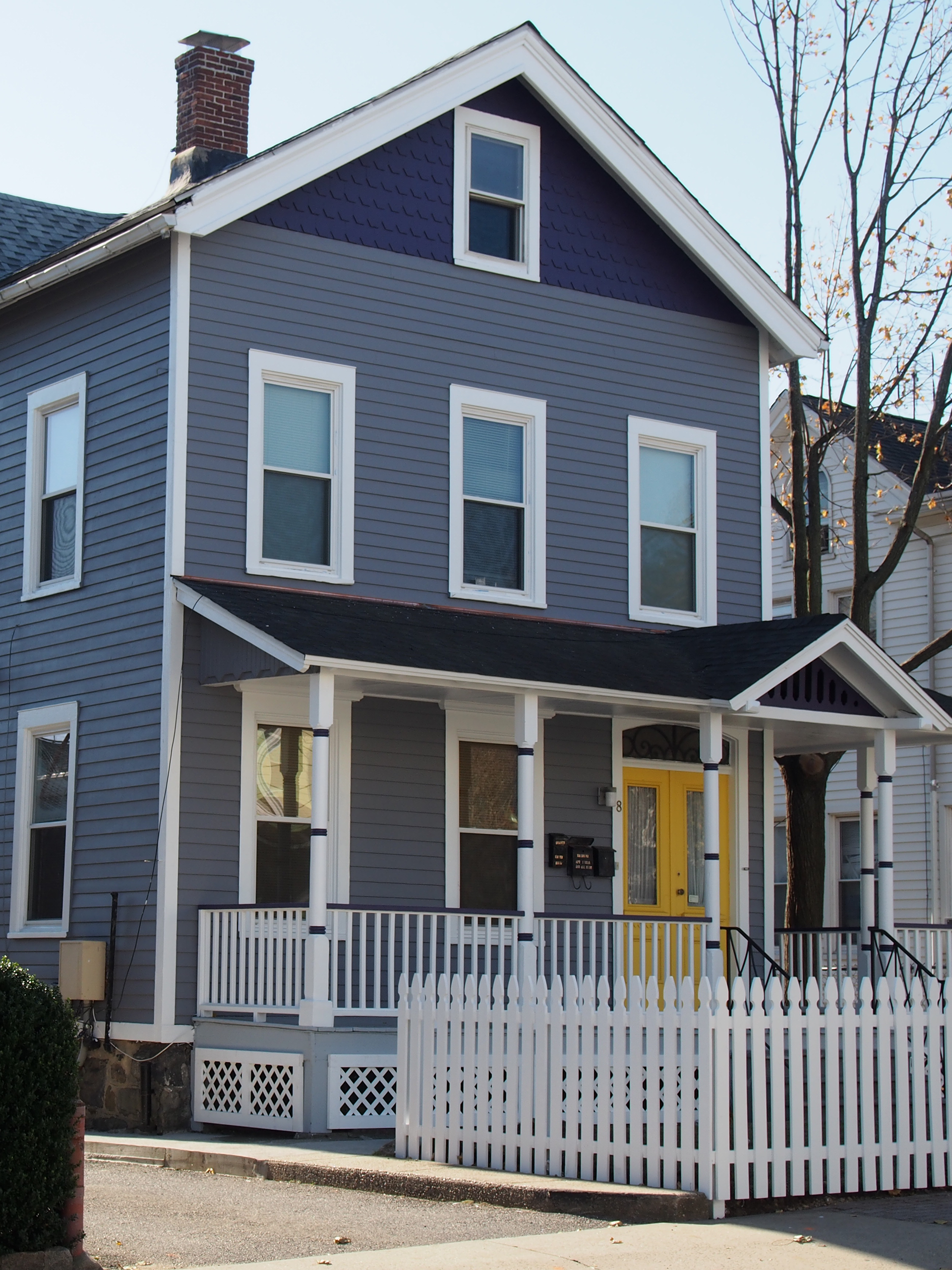 A two-story blue house with white trim, a yellow front door, and a white picket fence in front.