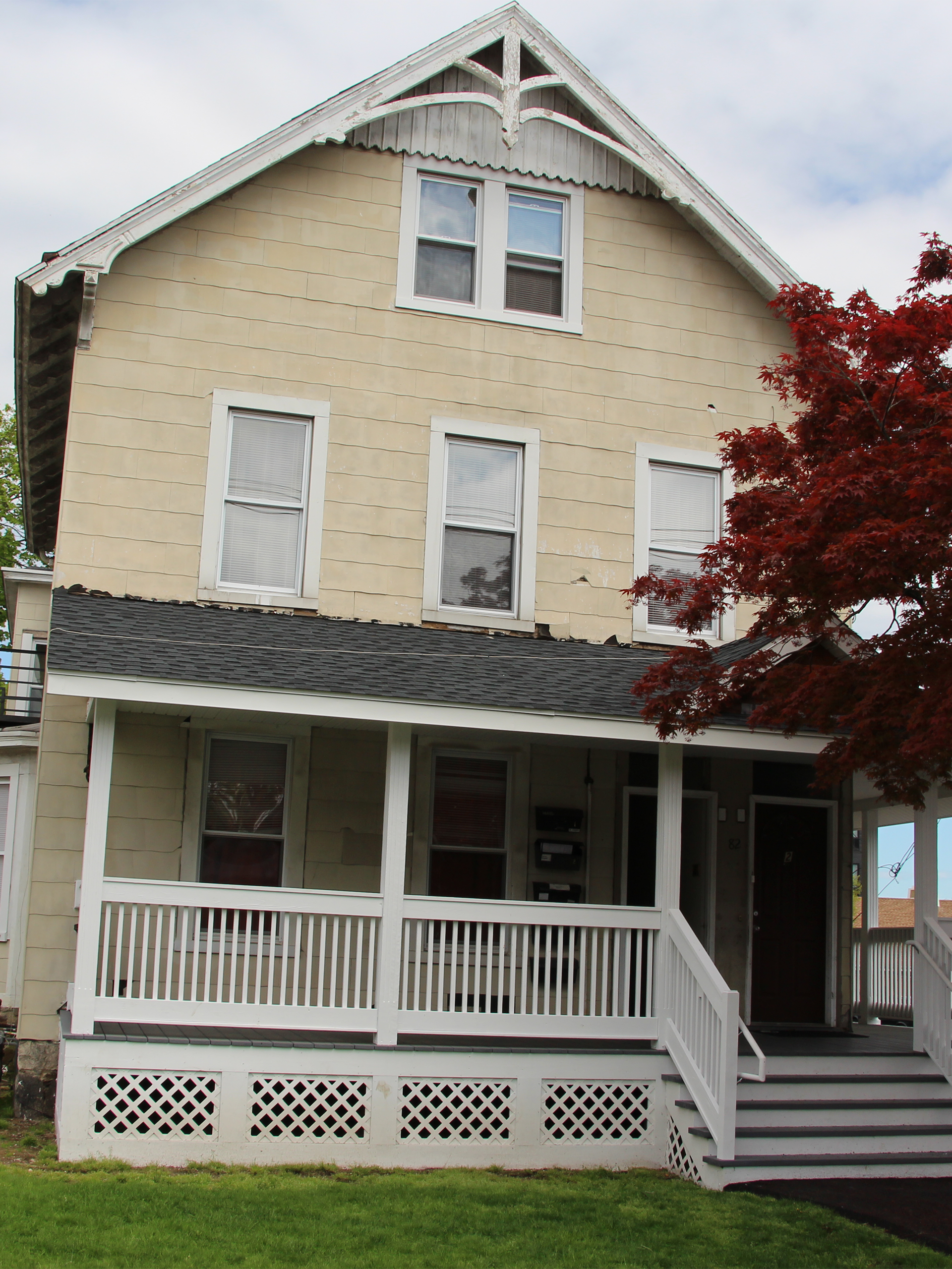 A three-story, light-colored Victorian house with a white front porch, lattice skirting, and decorative eaves.