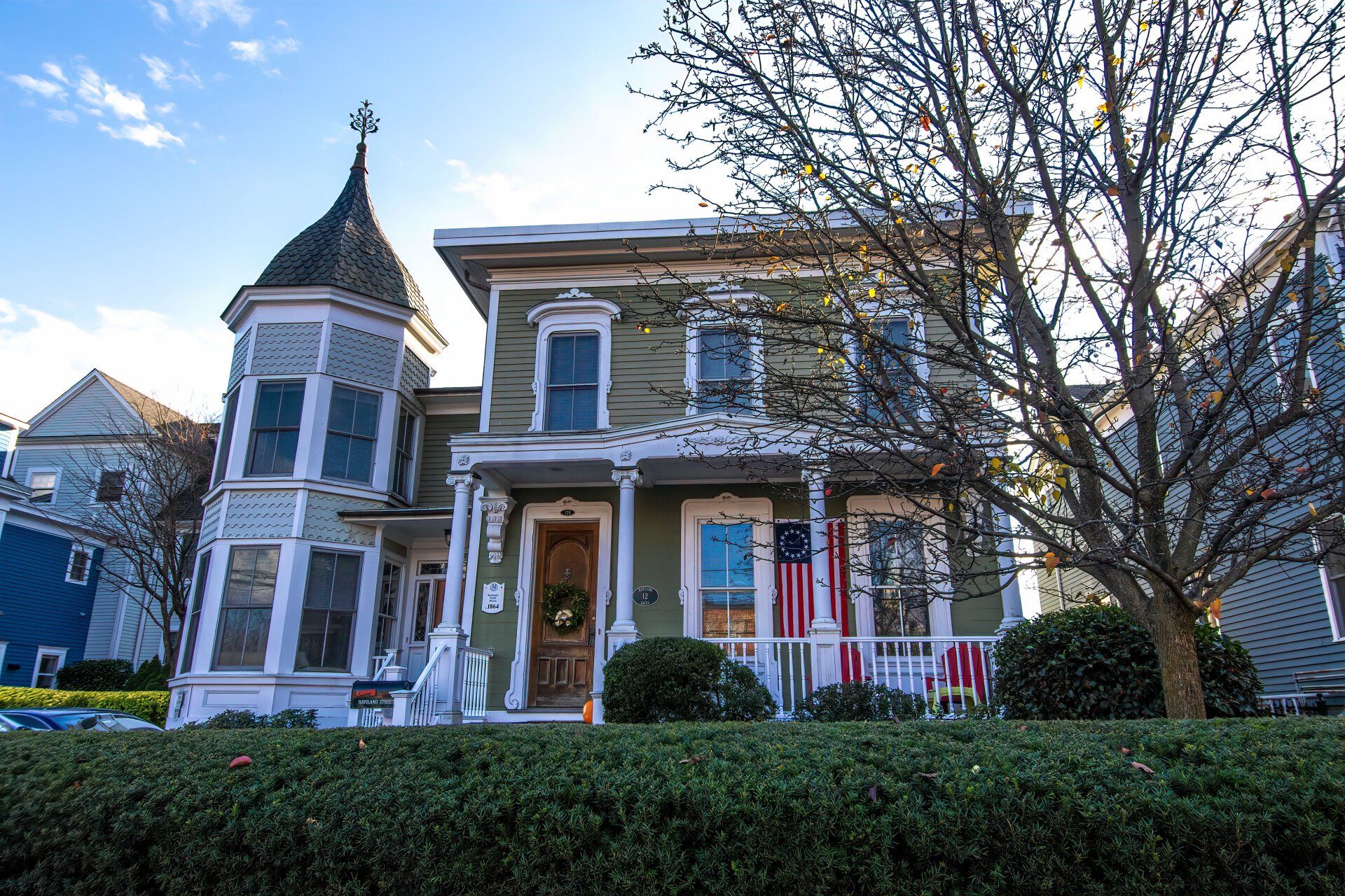 An olive-green Victorian house with a corner turret, ornate white trim, a front porch, and an American flag.