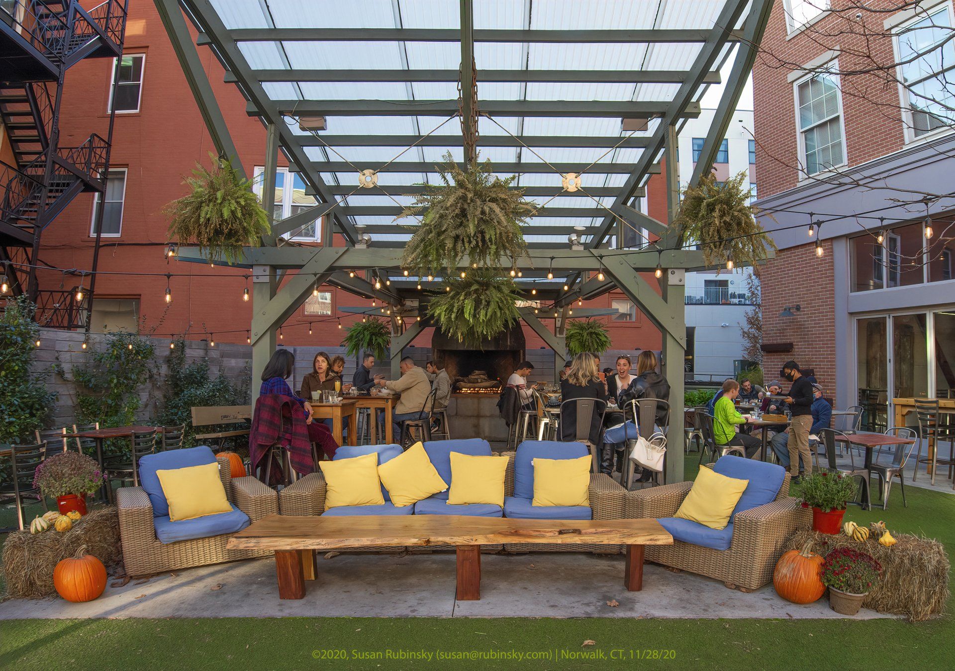 Outdoor patio with a wood-framed pergola, seating with yellow cushions, and hanging plants, decorated for autumn.
