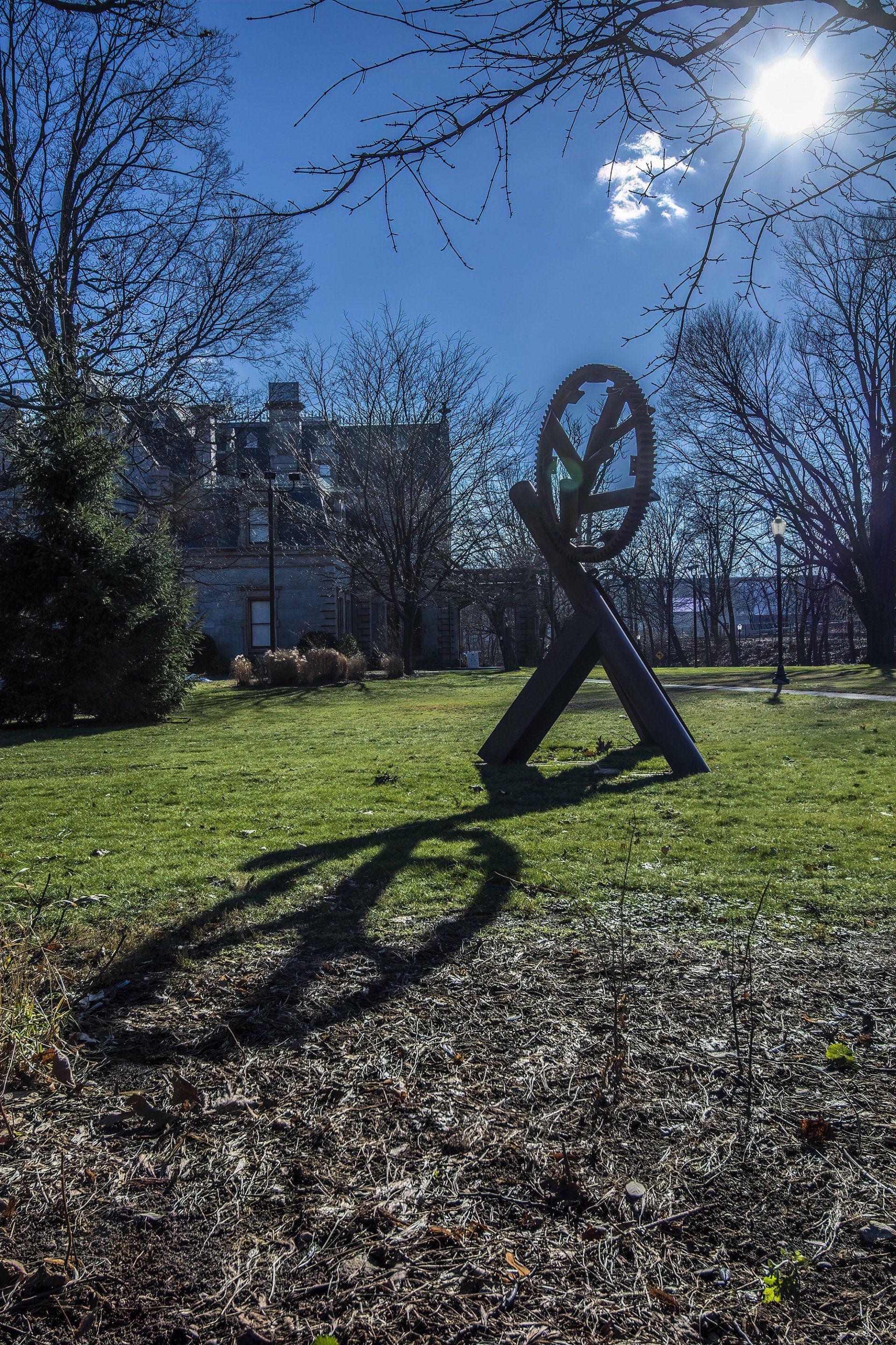 A dark, abstract metal sculpture of a peace sign stands in a grassy park on a sunny day, casting a long, distinct shadow.