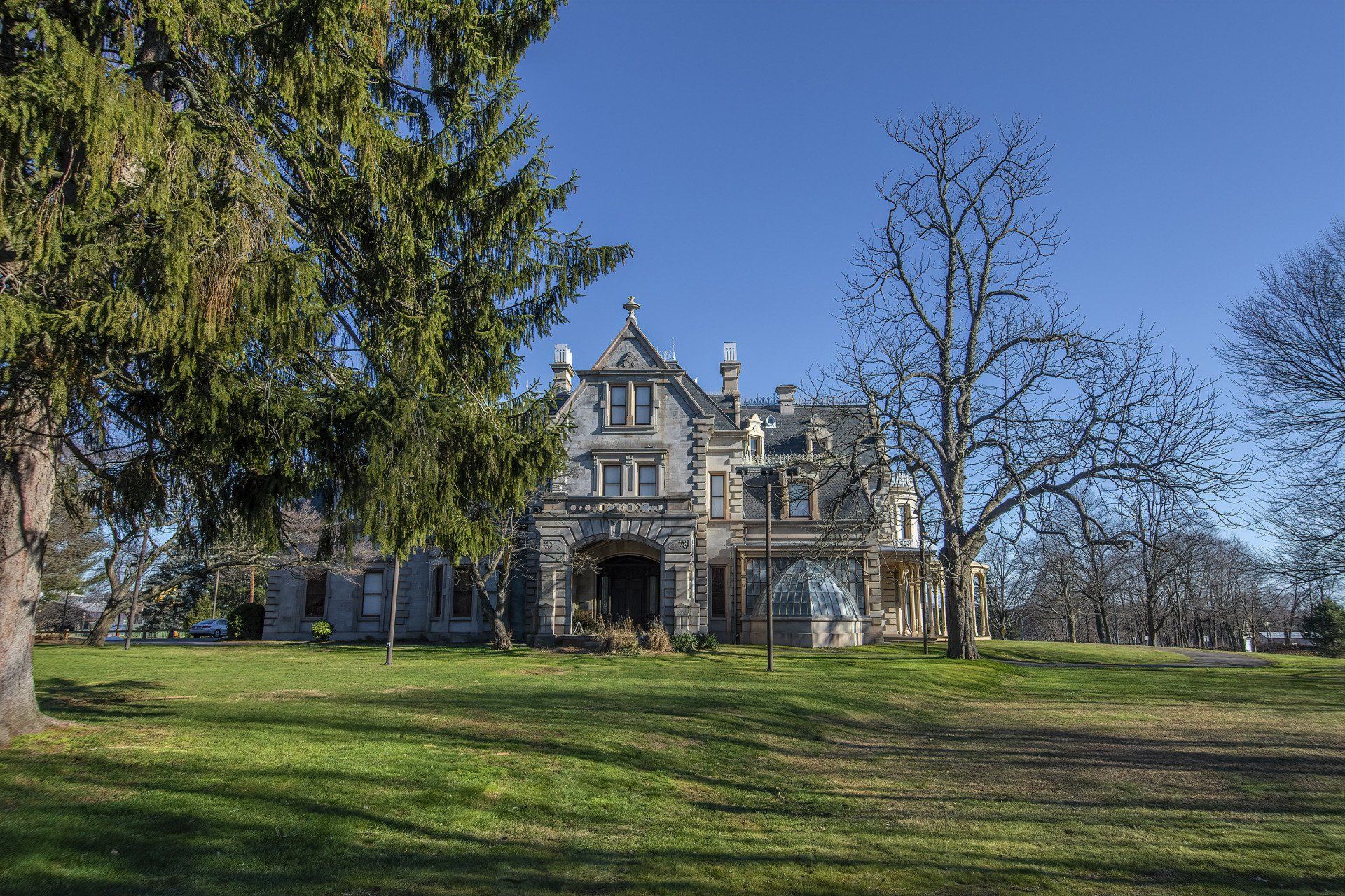 A grand stone mansion with a gabled roof sits on a grassy lawn under a clear blue sky, framed by trees.