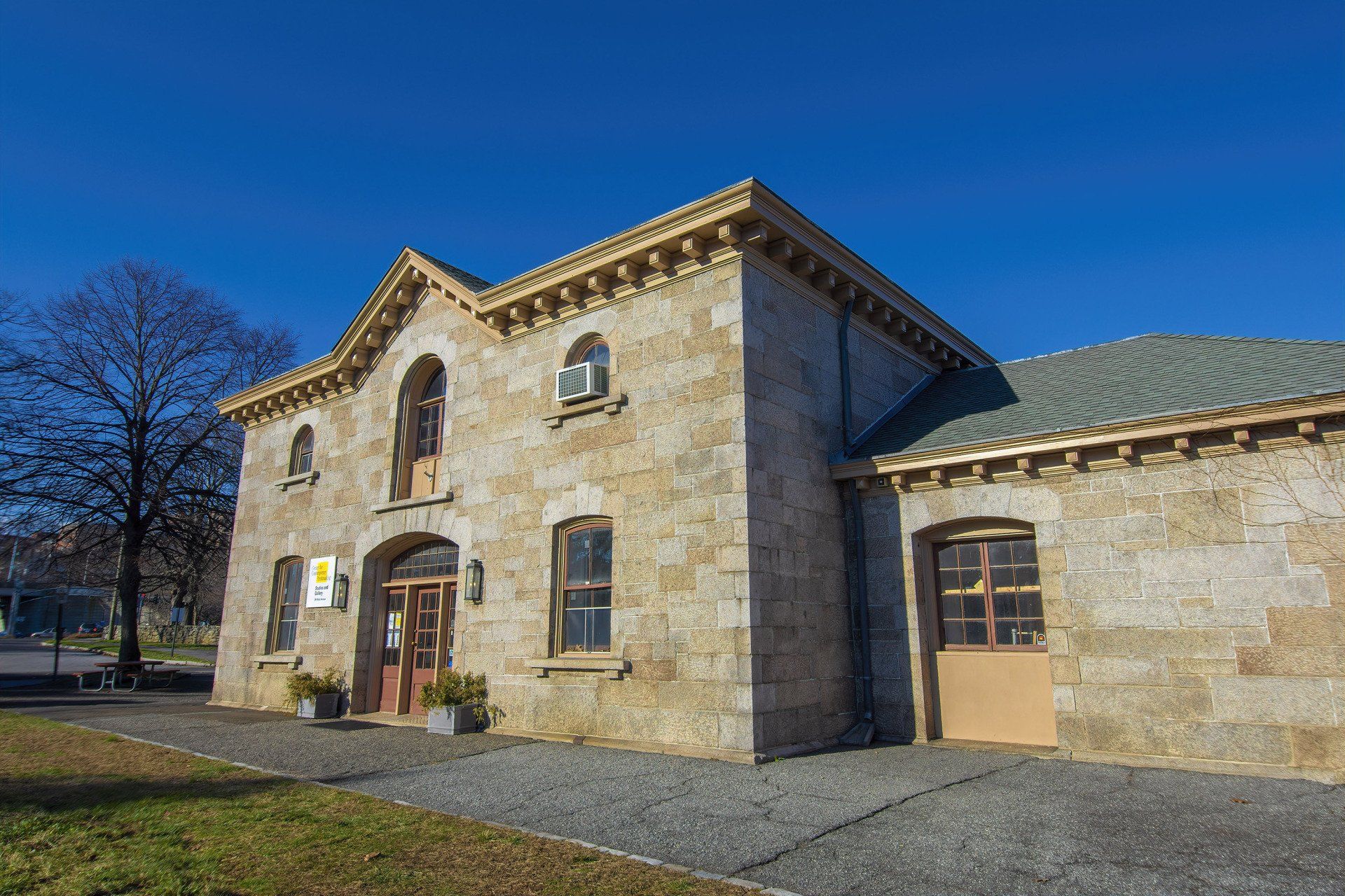 A stone building with a central gabled entrance, arched doorways, and windows under a bright blue sky.