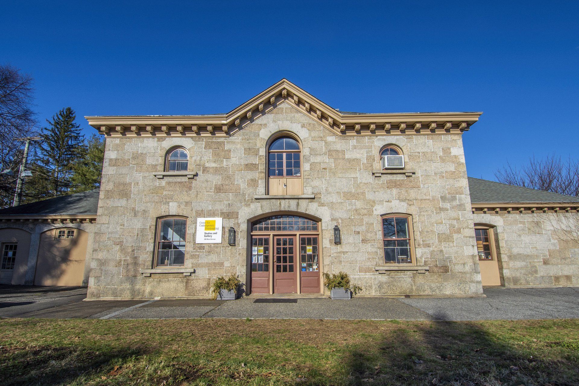 A historic, two-story stone building with a central gabled entrance, arched windows, and brown wooden doors against a sky.
