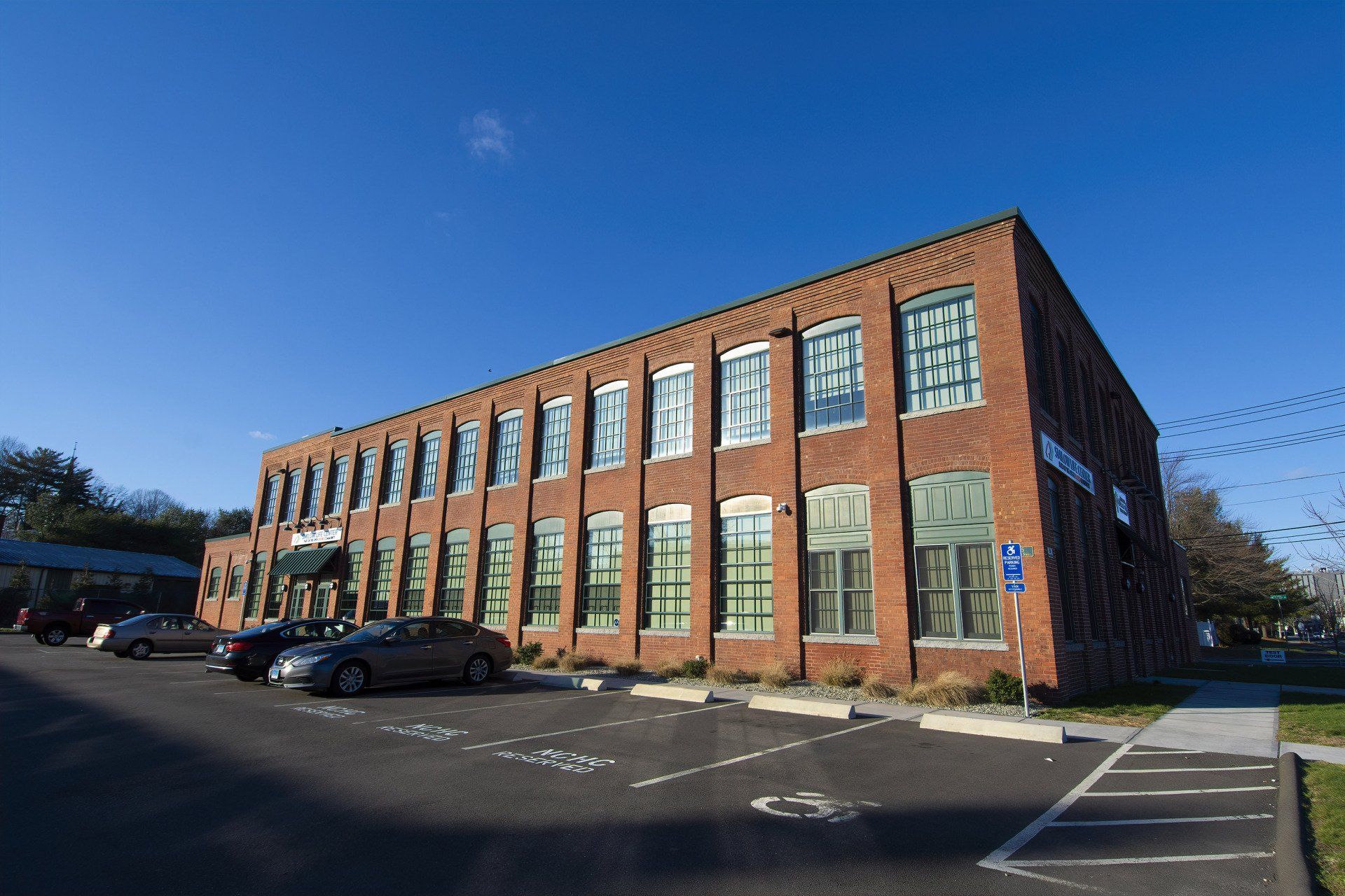 A three-story brick industrial building under a clear blue sky, with a parking lot in the foreground.