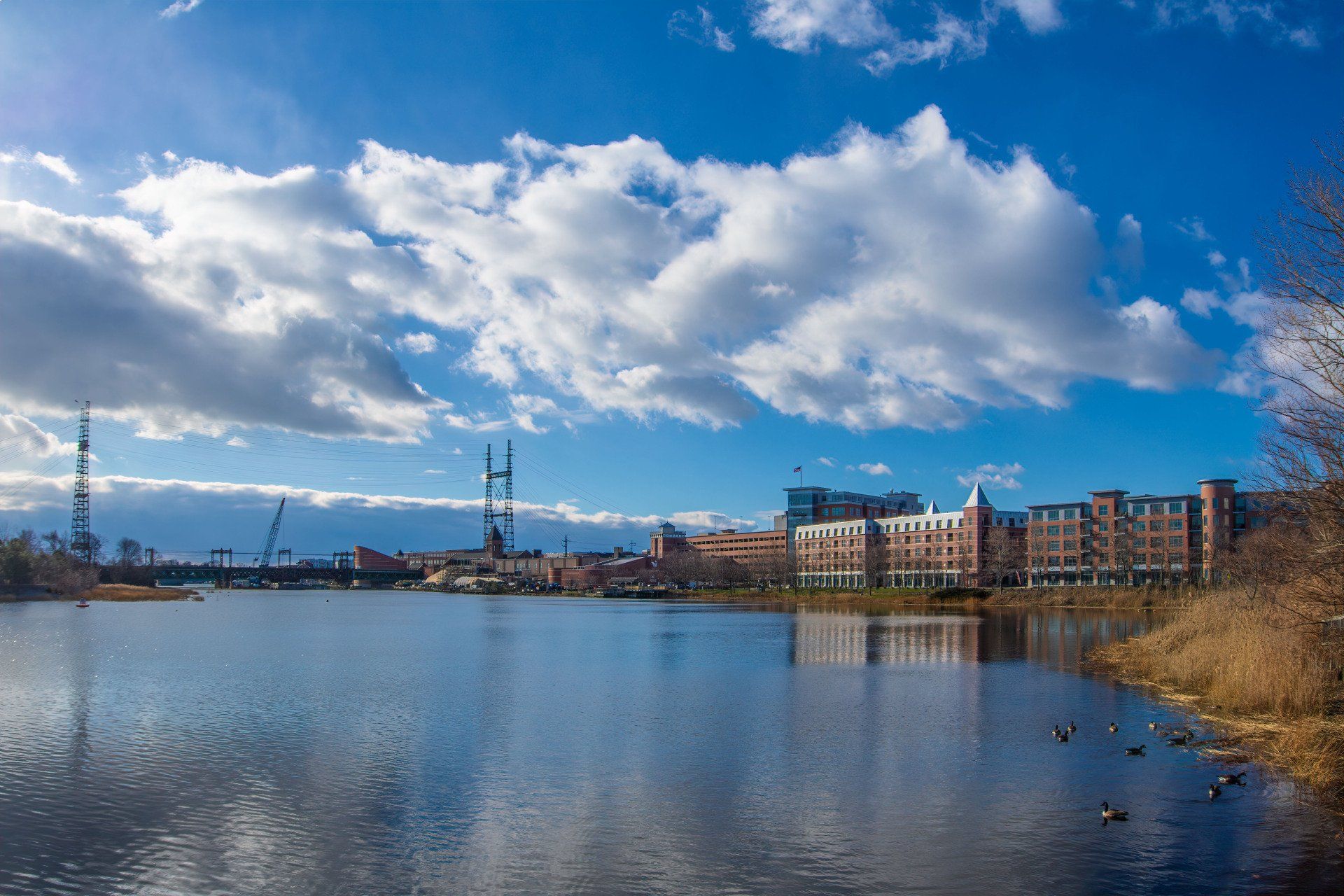 A calm river reflects a blue sky with white clouds, with buildings and construction cranes along the distant shore.