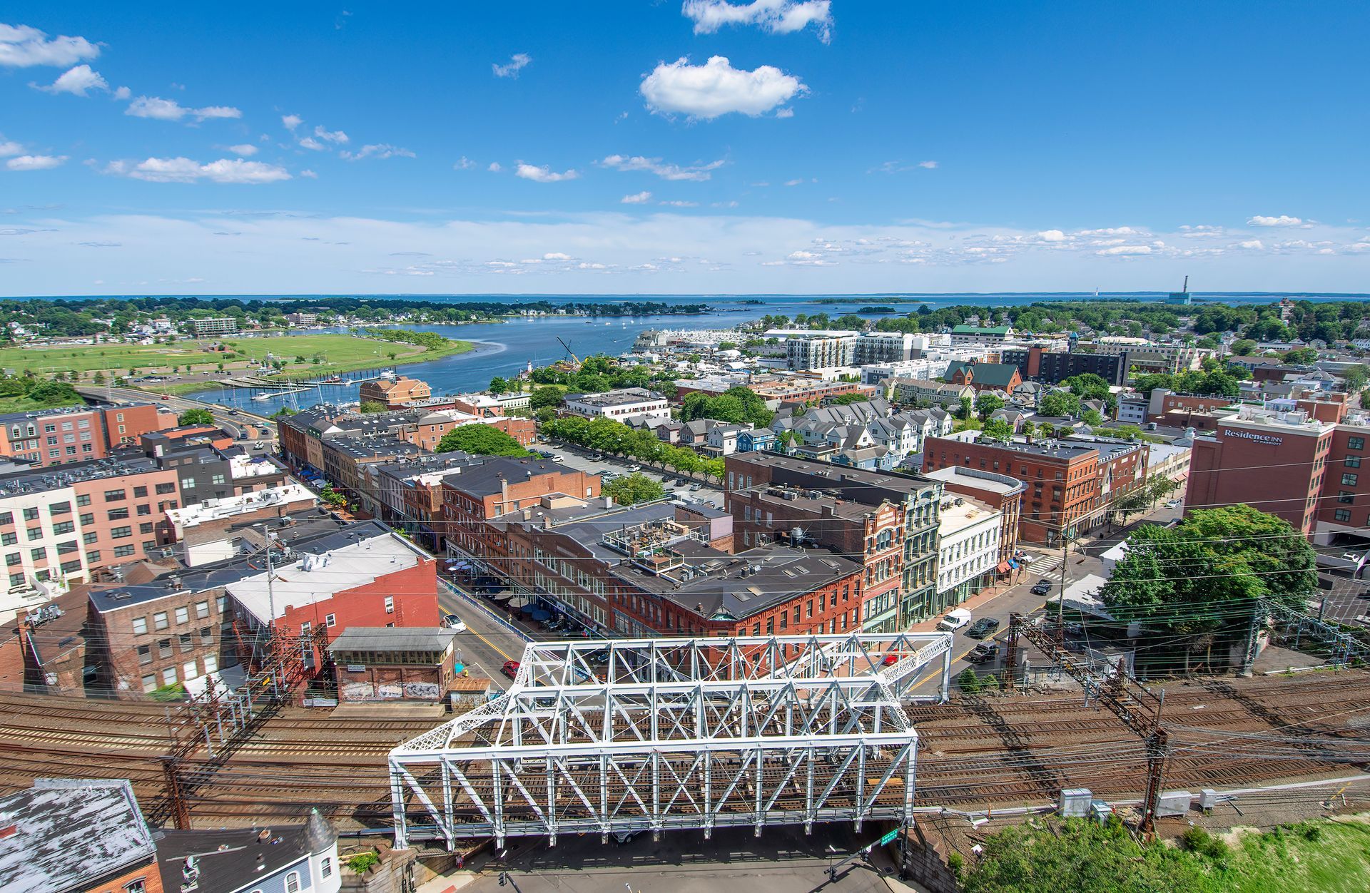 Aerial view of a coastal city, featuring a large white truss bridge spanning over railroad tracks near the harbor.
