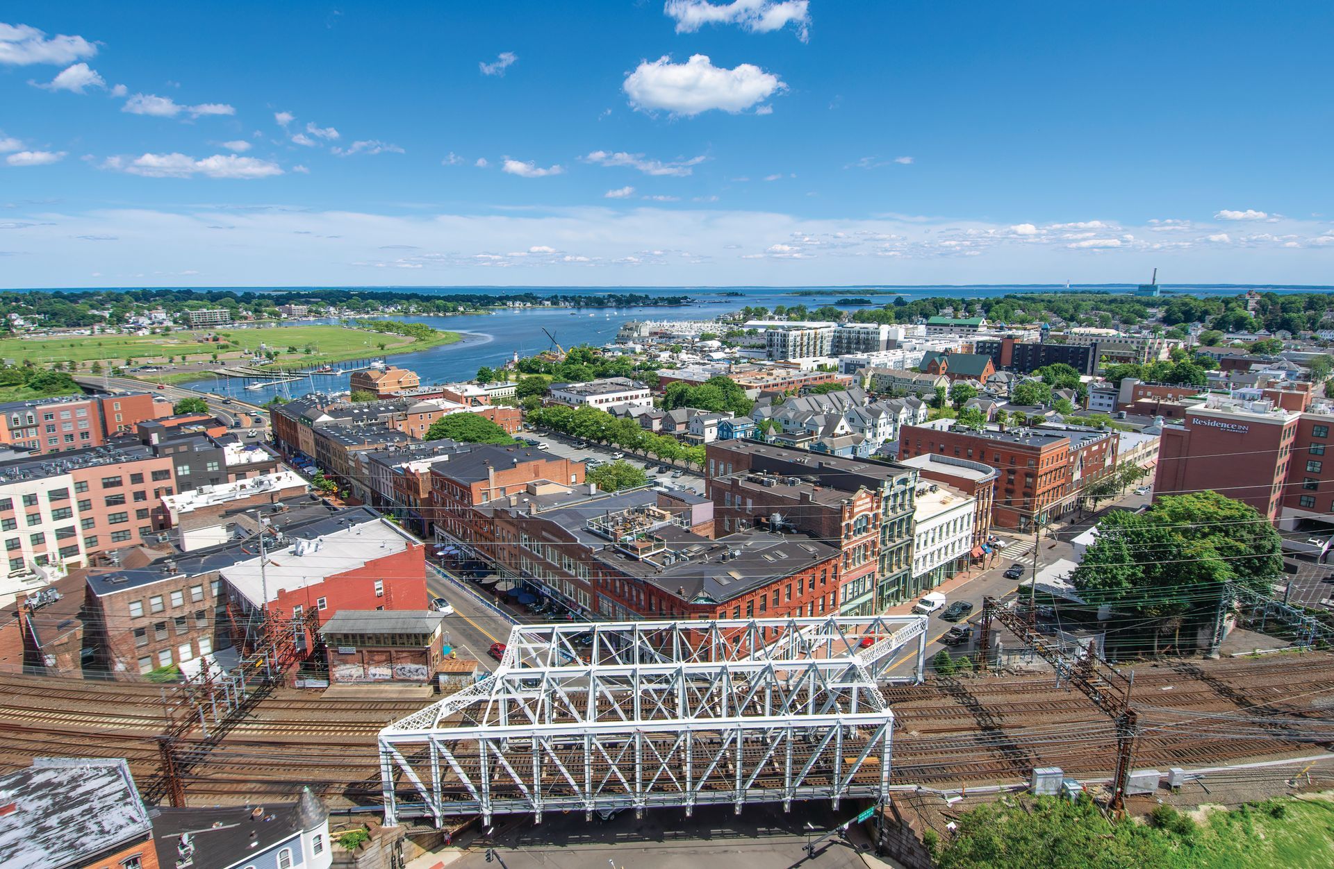 High-angle view of a city waterfront, featuring a large white railway bridge in the foreground and a blue harbor beyond.