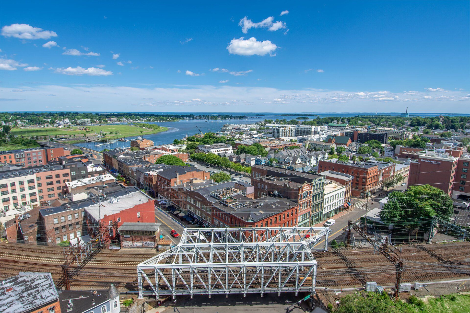 An elevated view of a city center with a white truss bridge spanning train tracks in the foreground and a waterfront bay.