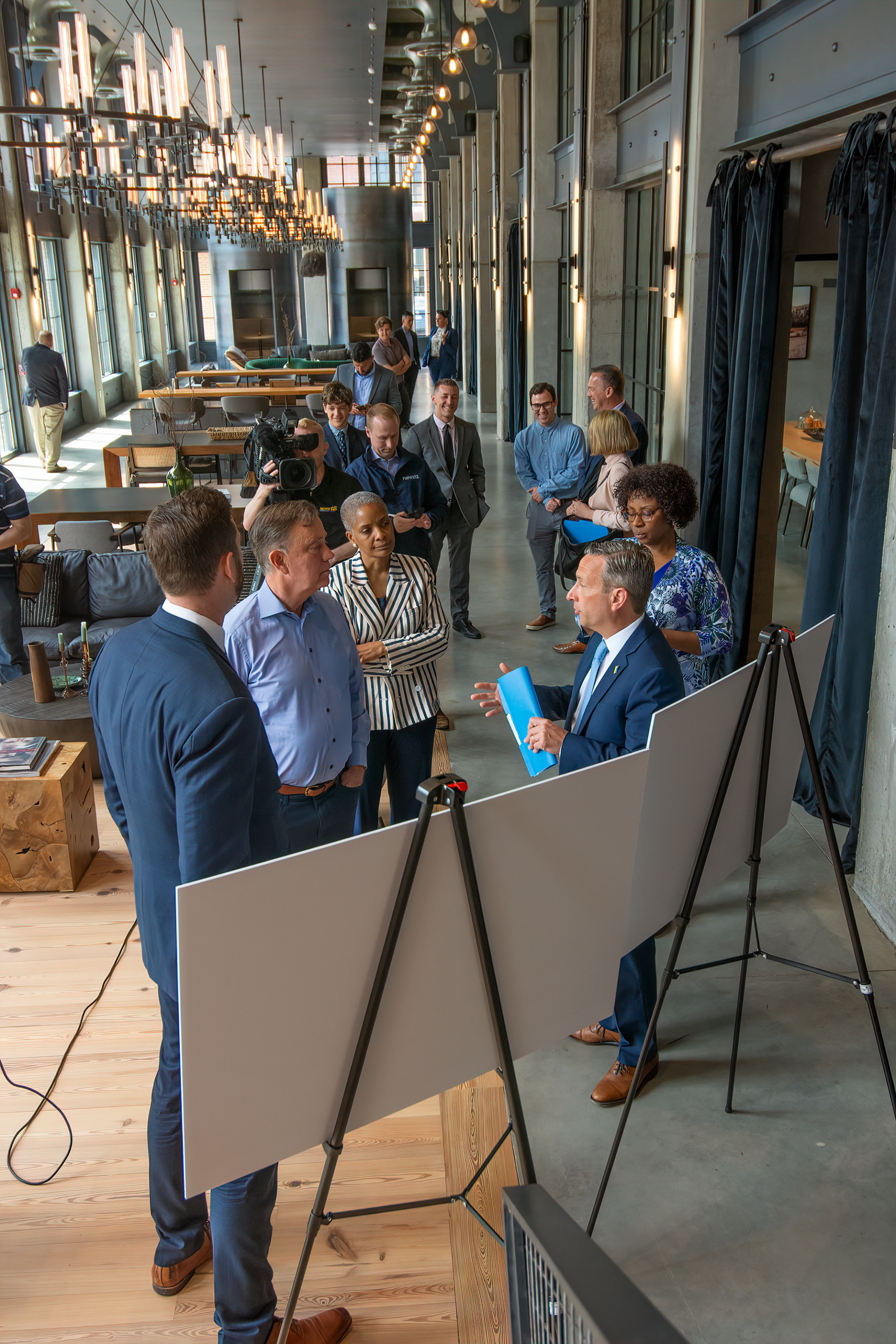 A group of people gathers in a modern, chandelier-lit hall as a man in a blue suit presents information on an easel.
