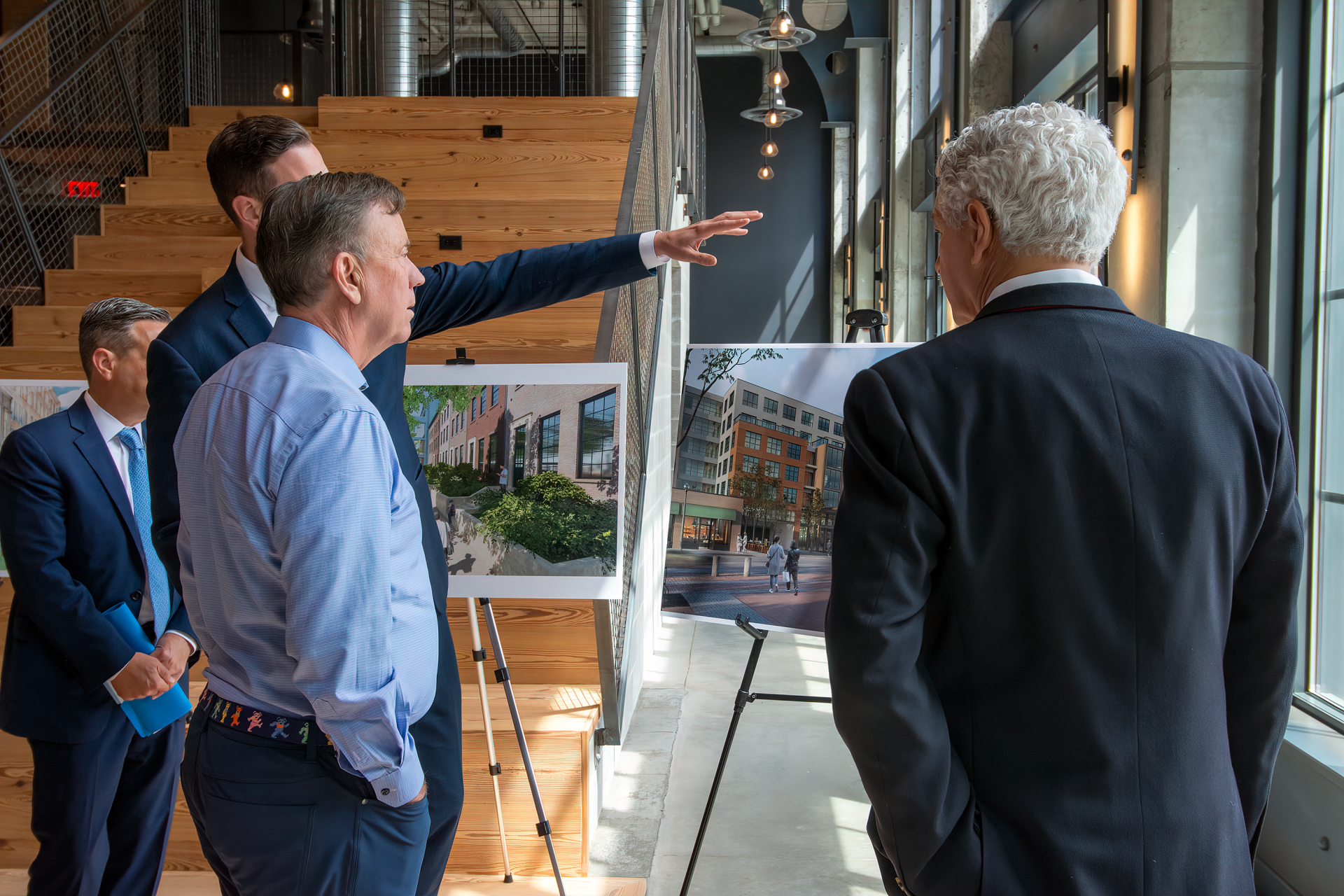 Four professionals in business attire stand in an office atrium, discussing architectural renderings on easels.