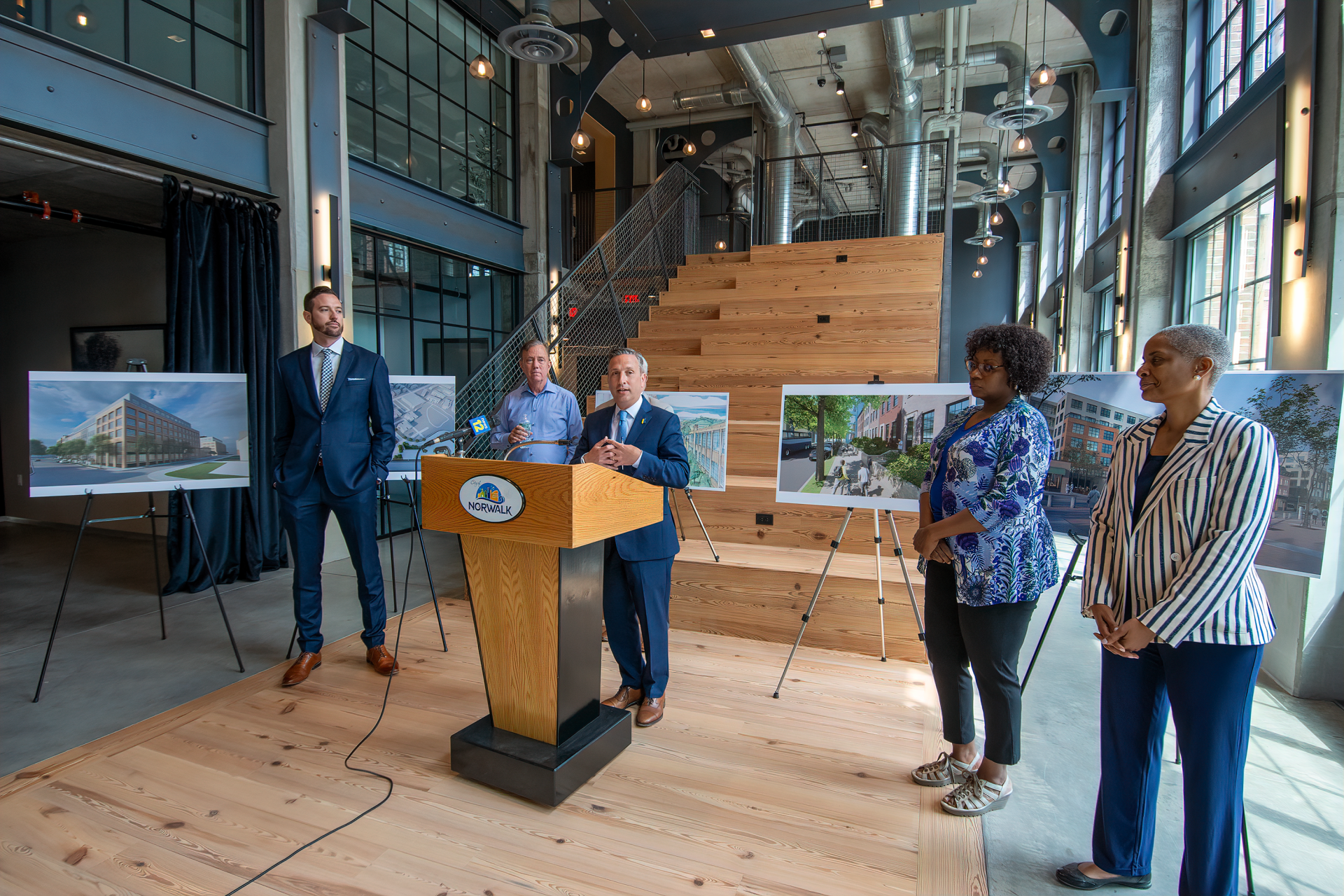 A group of five people stand on a wooden platform in an industrial room while one person speaks at a podium with displays.
