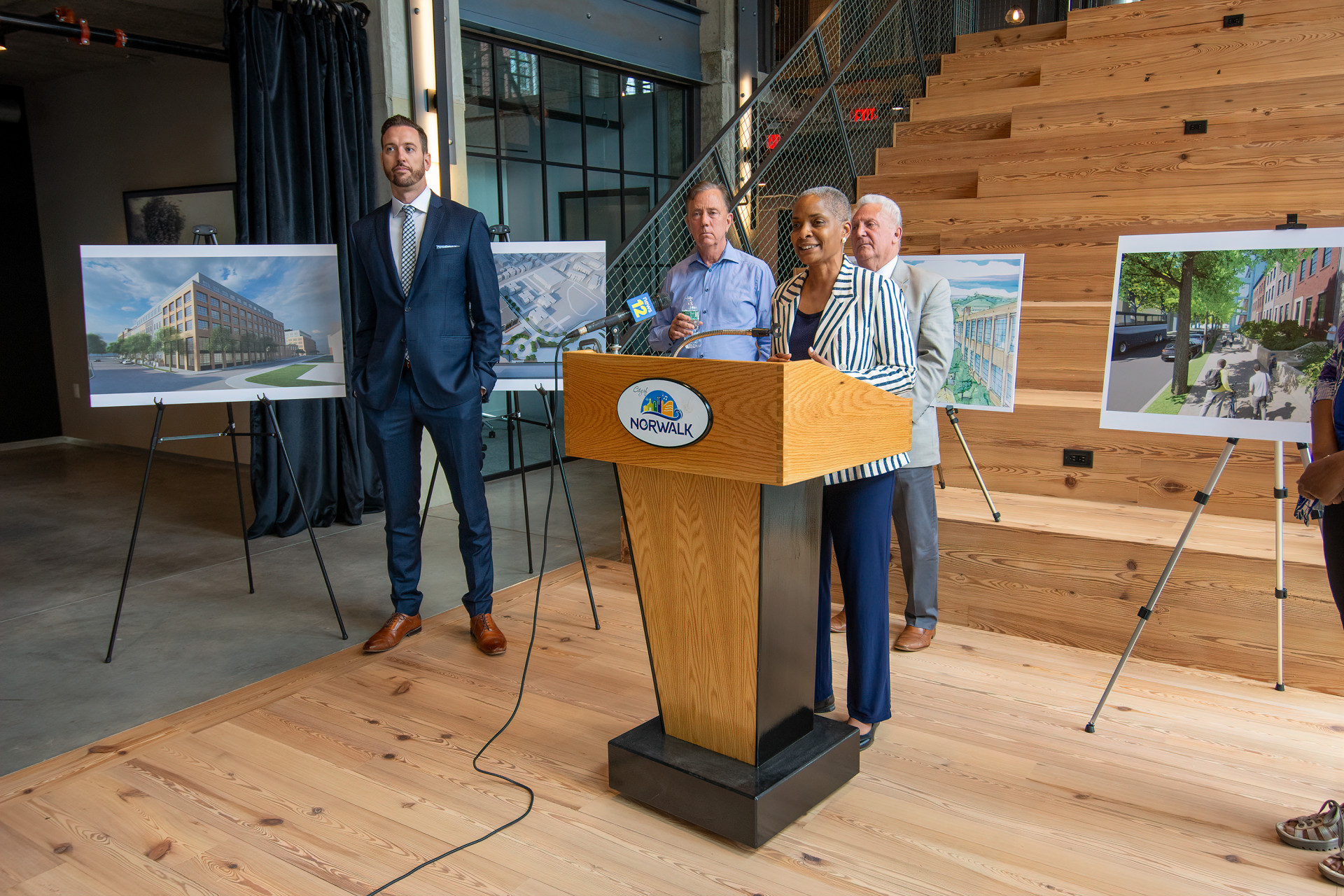 People stand behind a podium and architectural renderings in a modern, light-filled space with a wooden staircase.