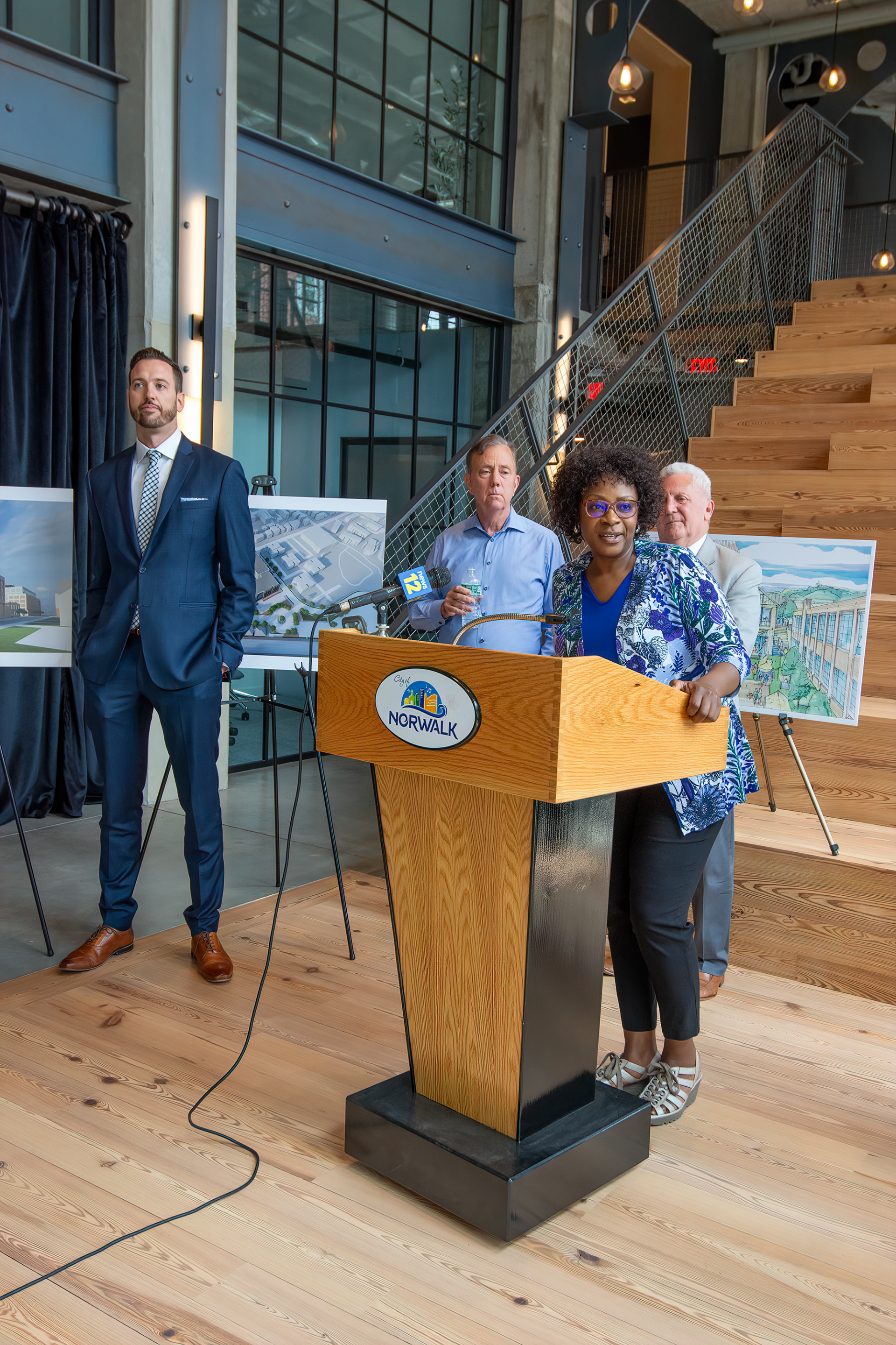 A group stands at a podium in a modern, multi-level building during an event, with project renderings displayed on easels.