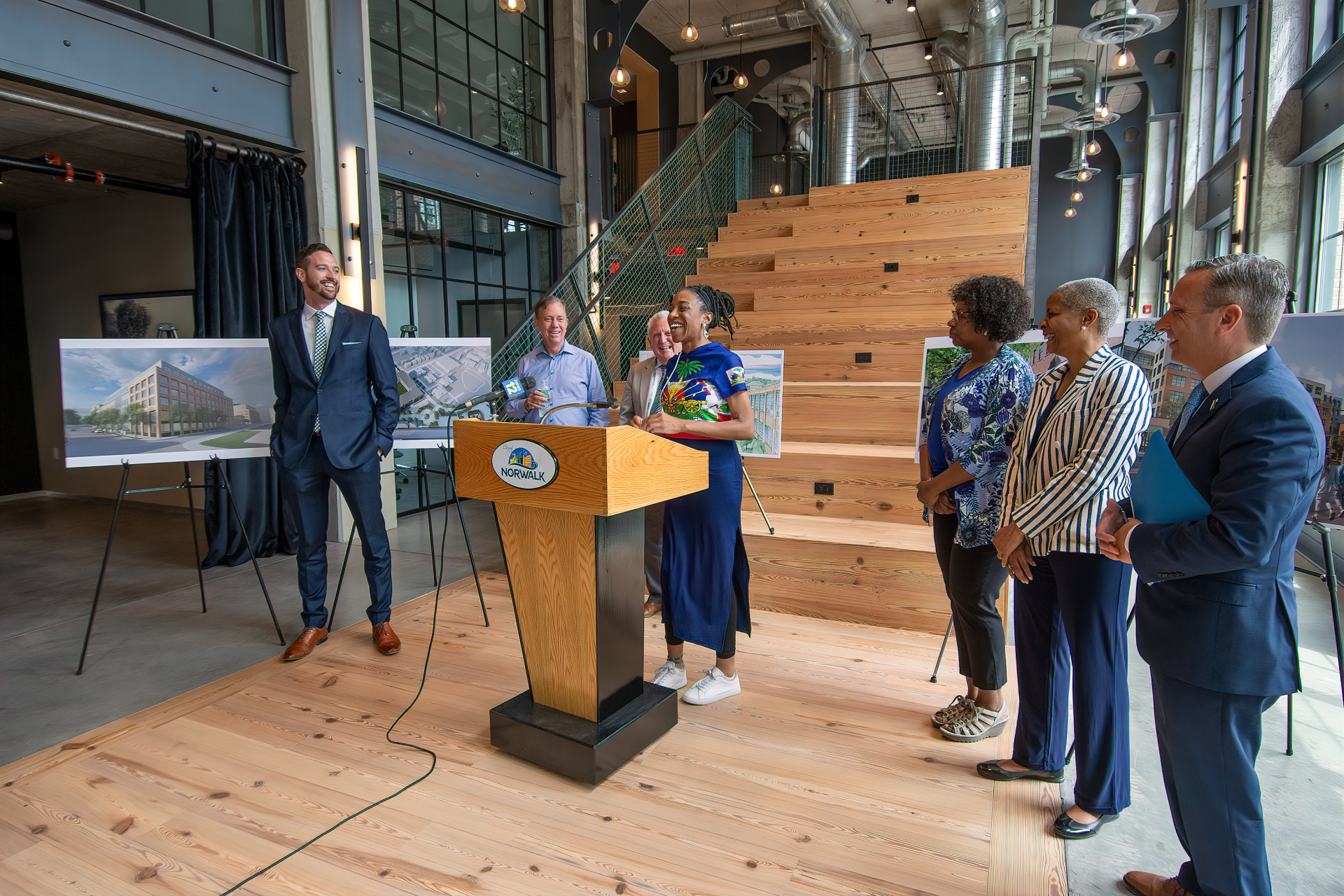 A group stands on a wooden stage by a lectern at an indoor event, with architectural renderings displayed on easels.