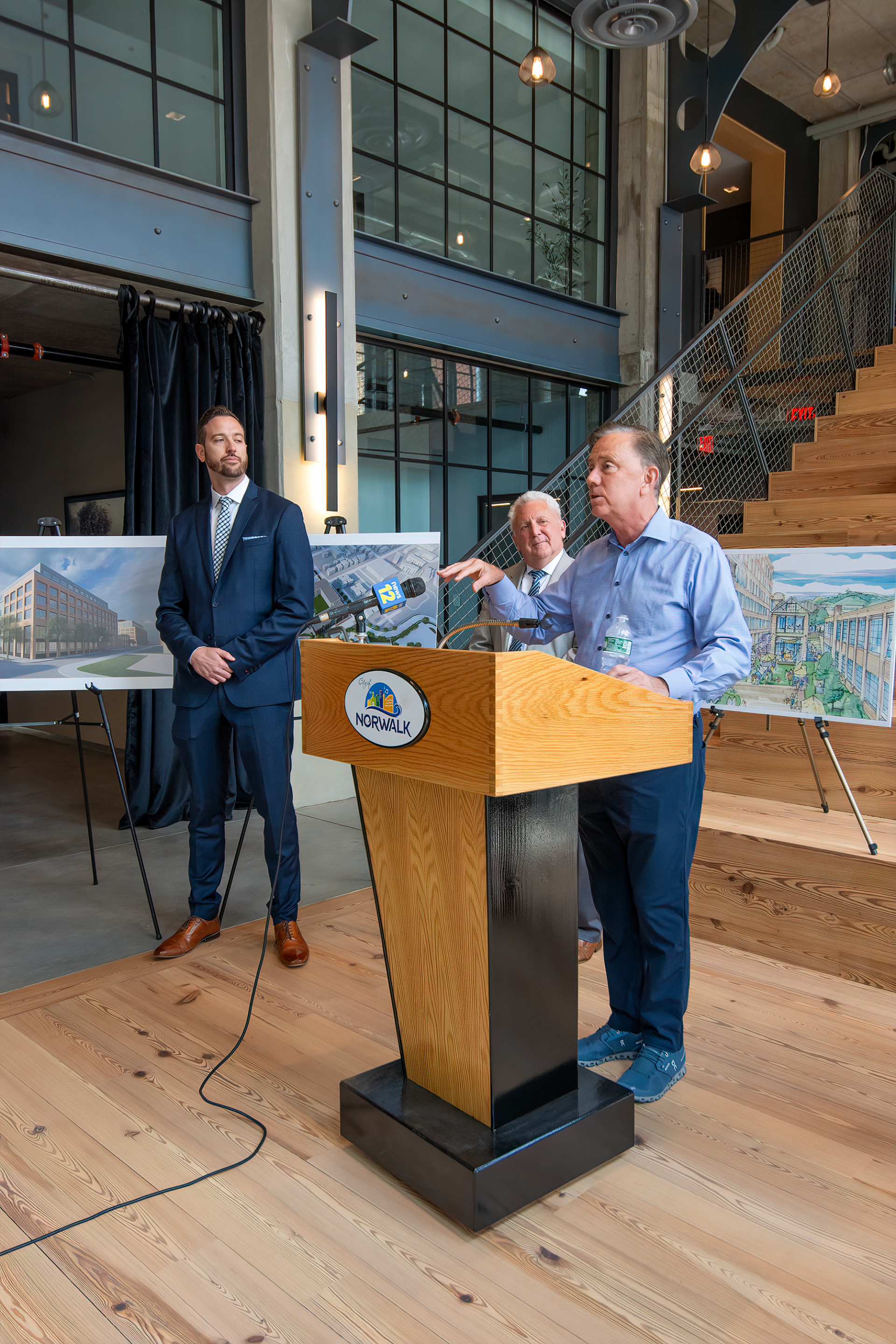 A speaker at a podium with two other people standing behind him in an industrial-style office space with architectural plans.