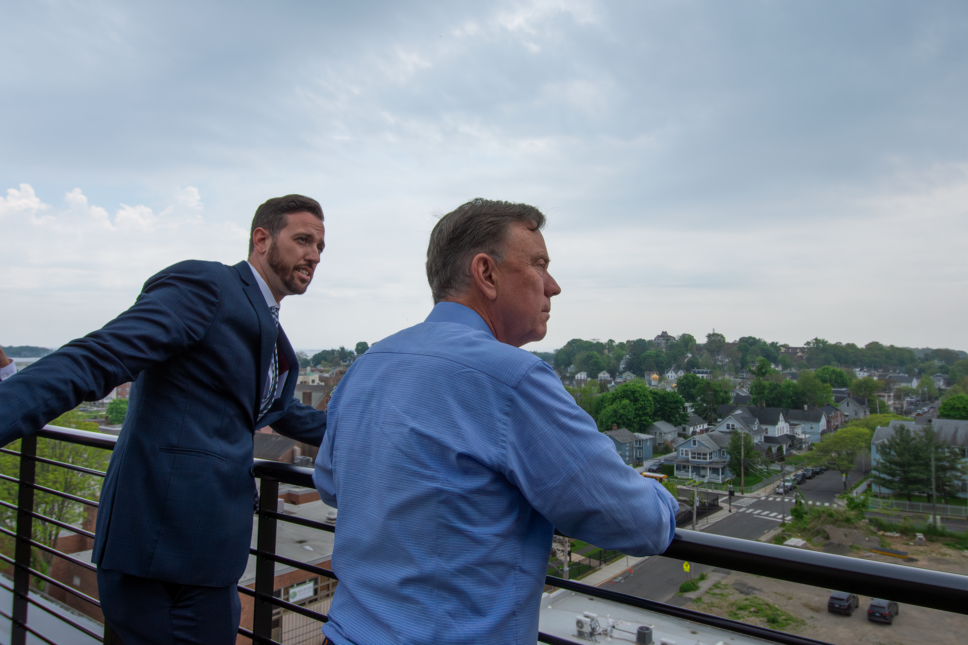 Two men standing on an outdoor balcony overlooking a suburban landscape under a cloudy sky.