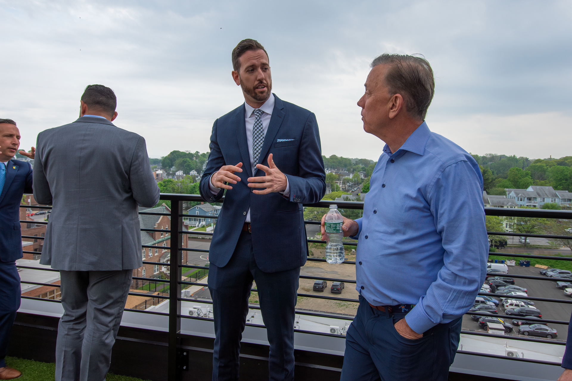 Three people in professional attire have a conversation on an outdoor rooftop terrace with a view of a town below.