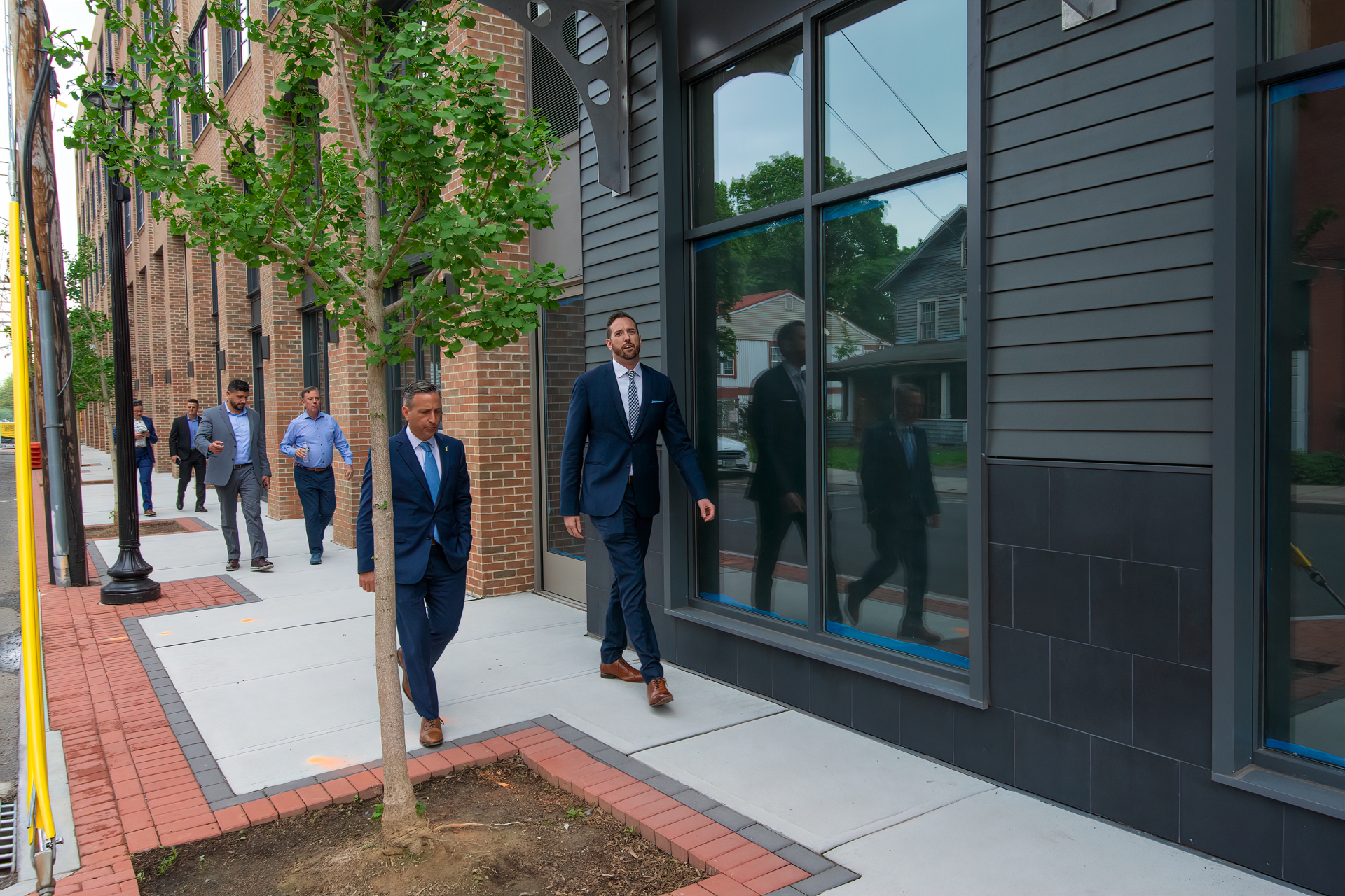 A group of people in business attire walking along a modern building sidewalk past a young tree.