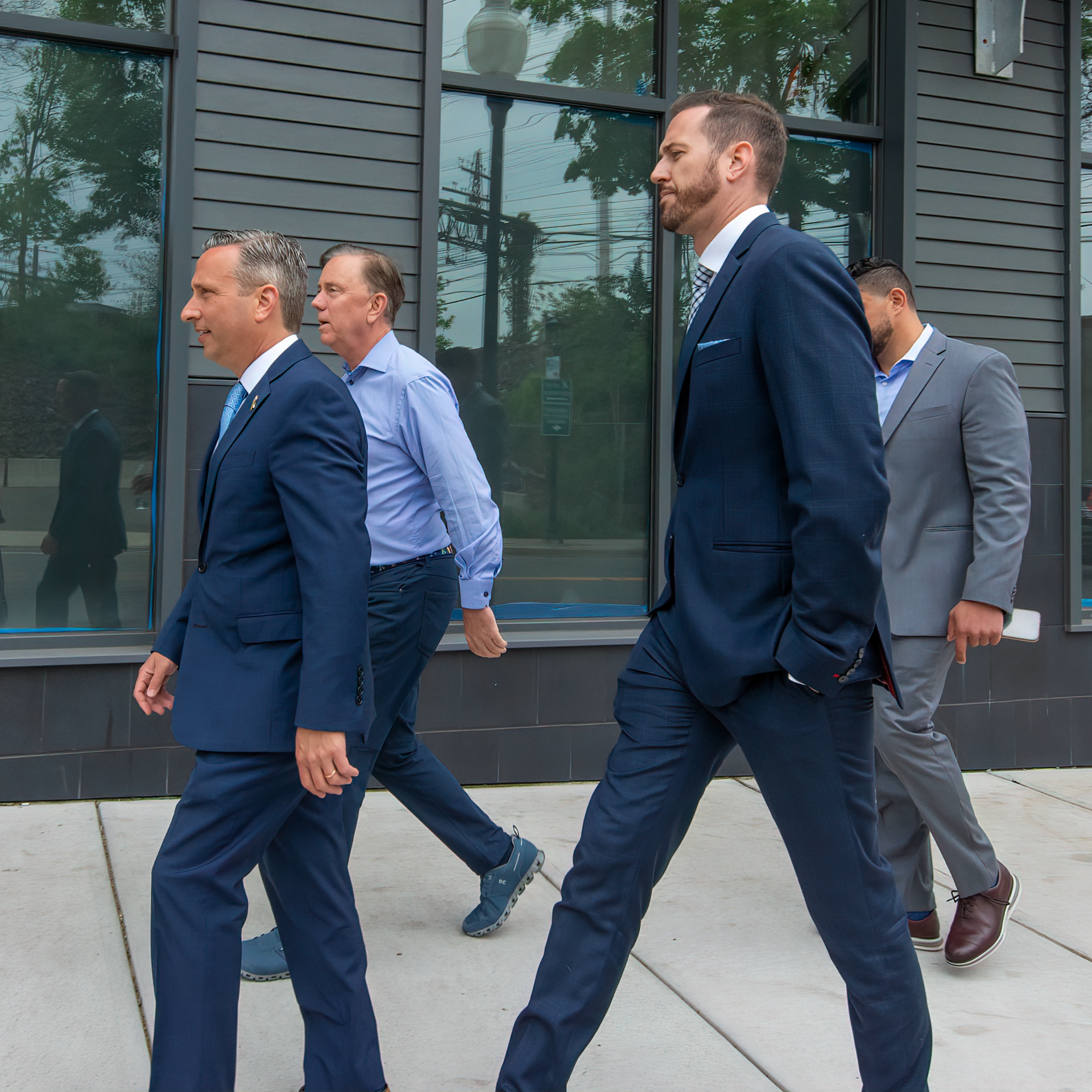 Four people in professional business attire walk in a line along a sidewalk past a dark building with large windows.