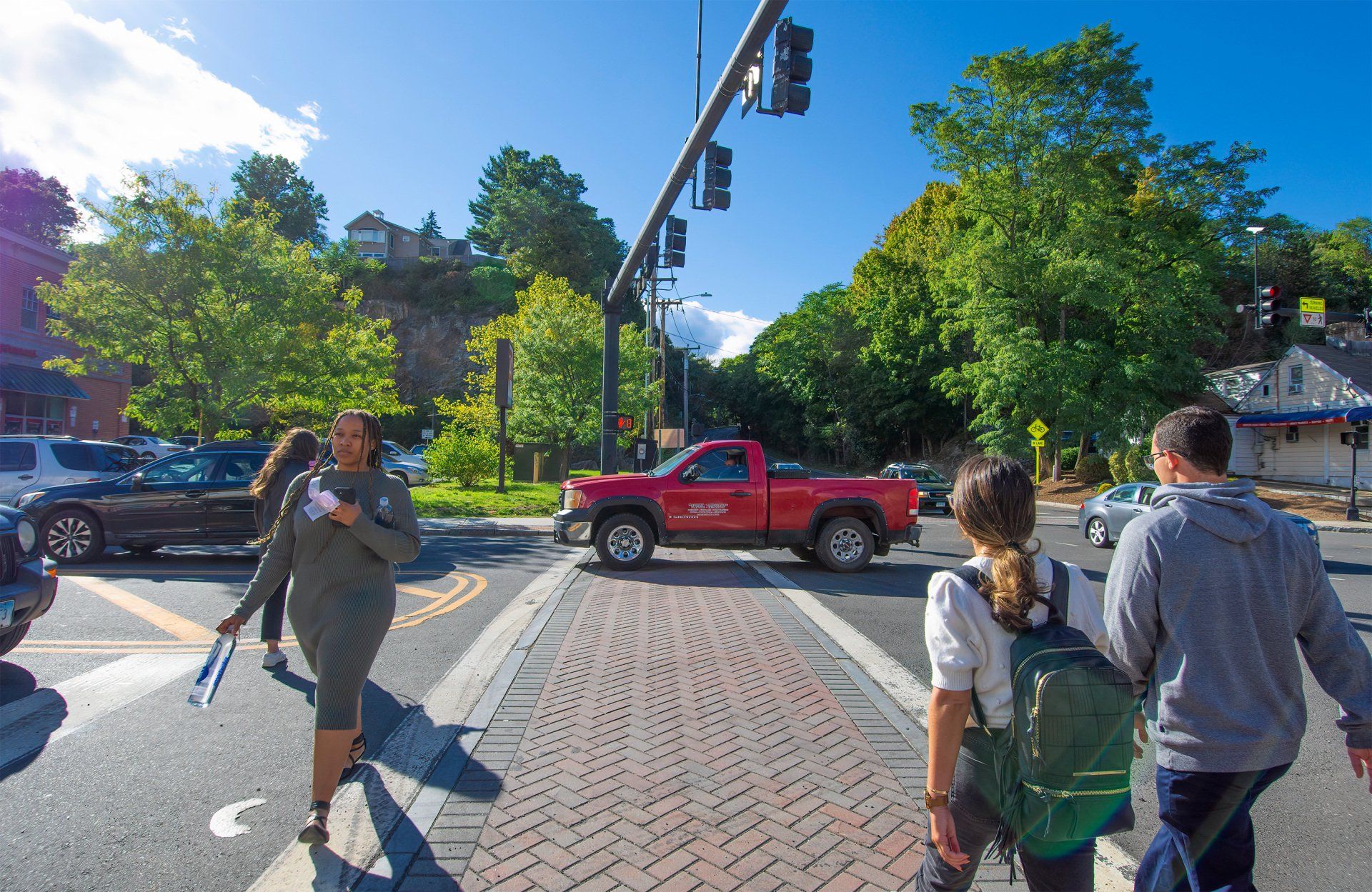 Pedestrians walk across a brick-paved crosswalk at a sunny intersection as a red truck drives past.
