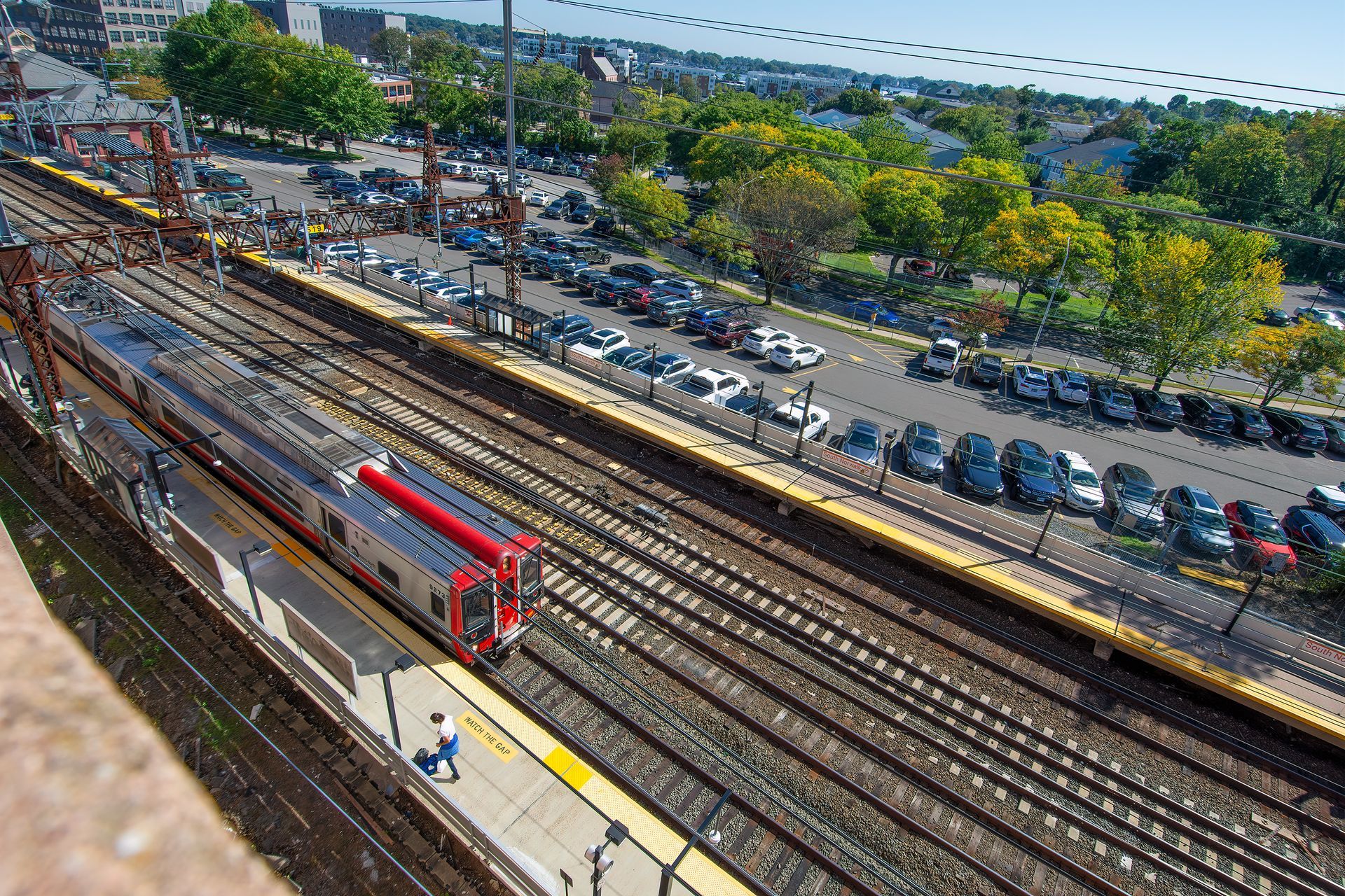 A high-angle view of a red and silver commuter train arriving at a station platform next to a large parking lot.
