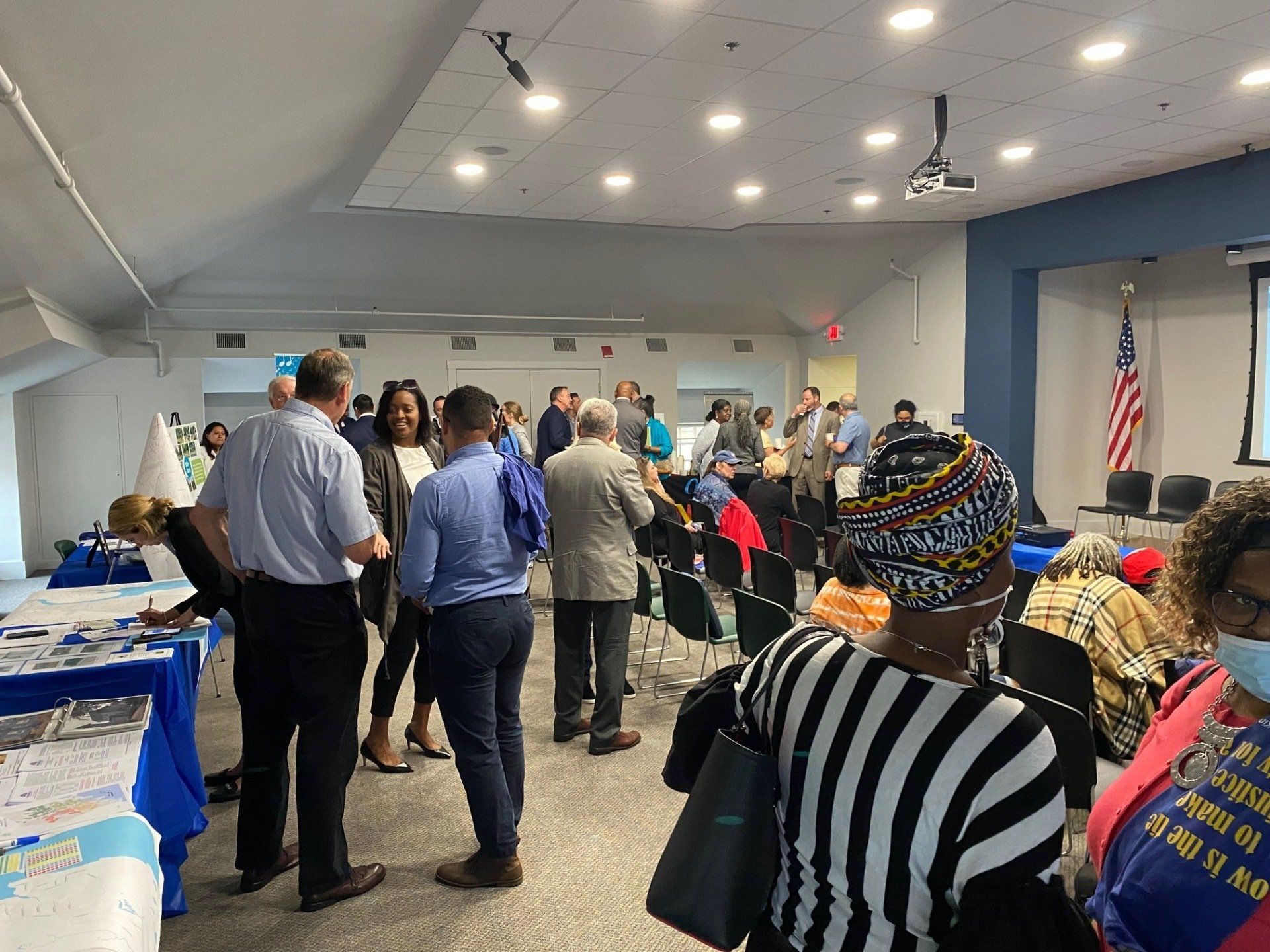 People gather for an indoor event, talking and mingling in a large room with tables, chairs, and an American flag.