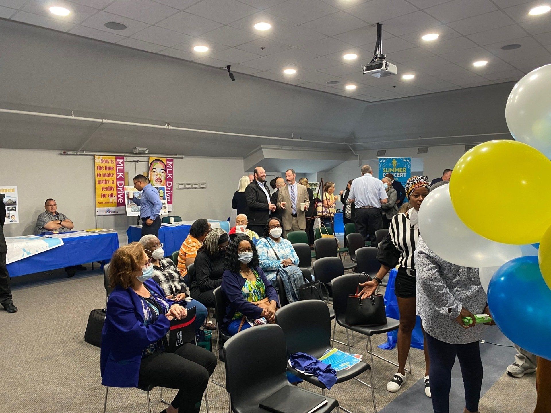 A group of people gathers in a room with blue-covered tables, posters, and balloons for an indoor social event.