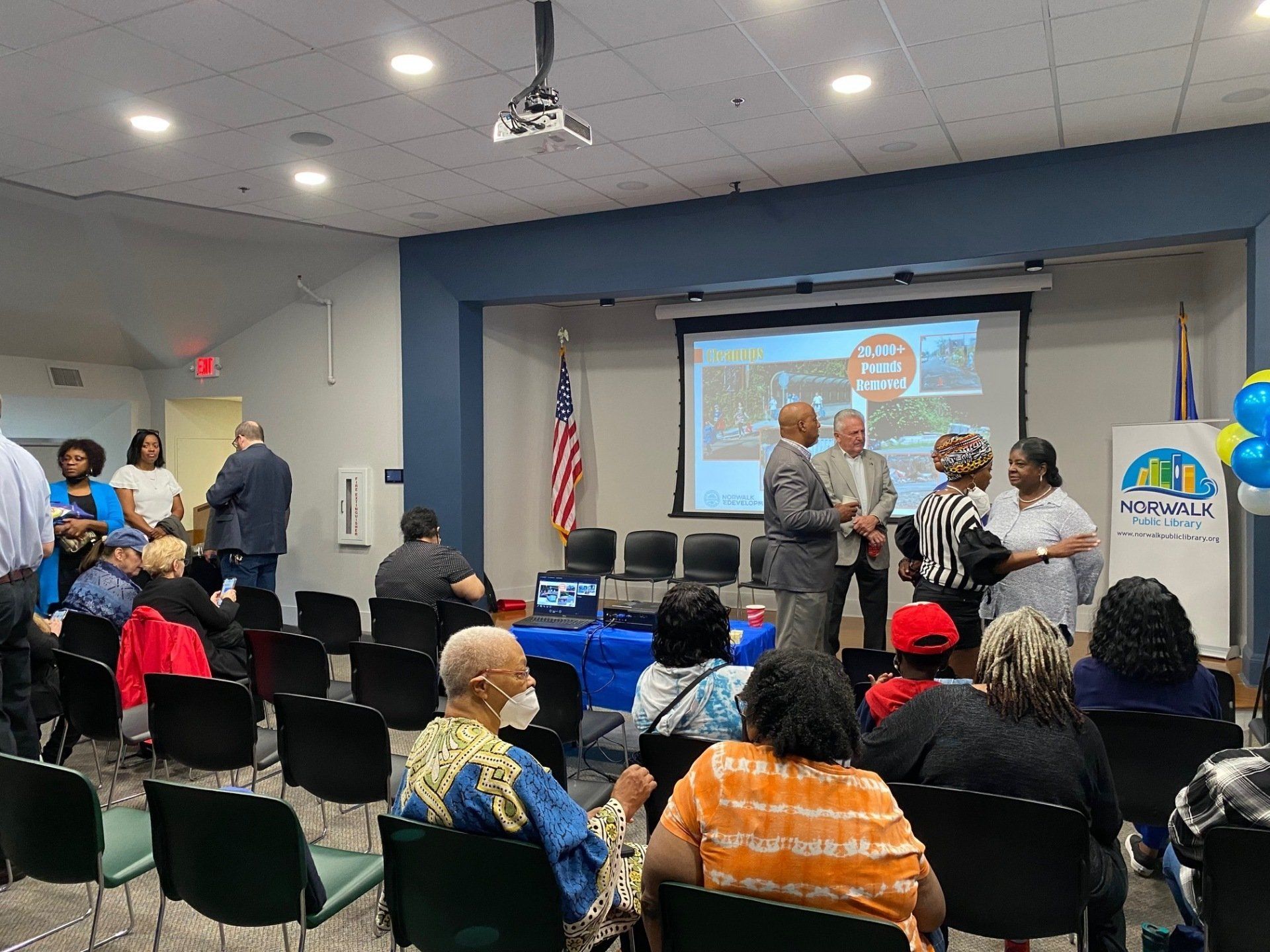 Attendees listen to a presentation in a room featuring a large projection screen, an American flag, and a banner.