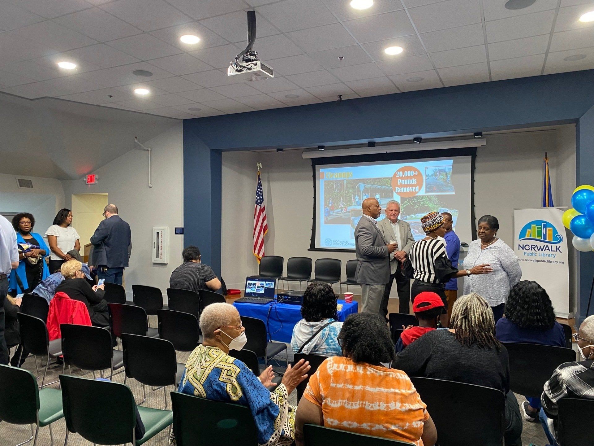 A group of people listens to a presentation in a meeting room with a projected screen, American flag, and event signage.
