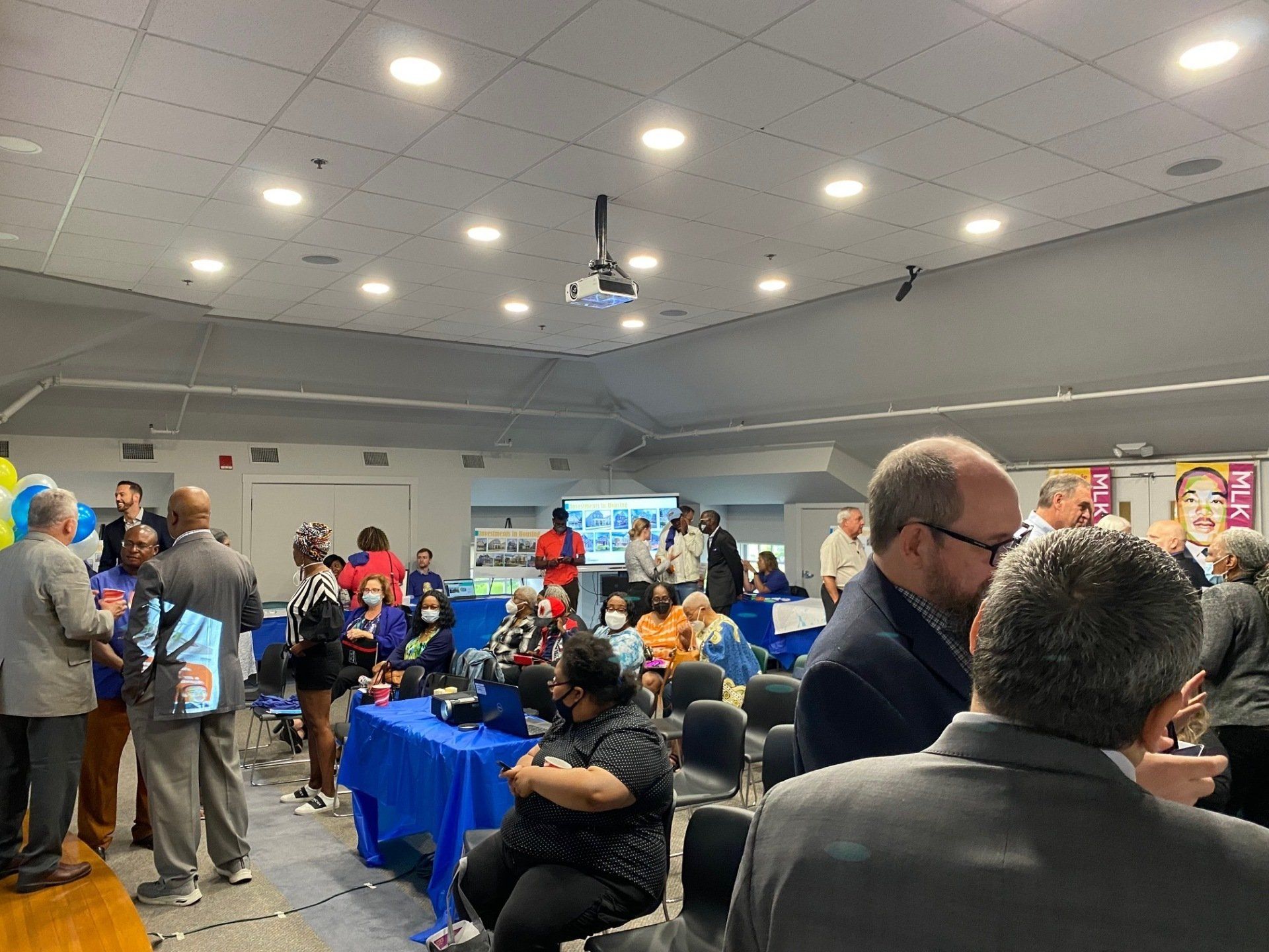 A large group of people gathers in a well-lit community hall with blue-clothed tables, appearing to attend an event.