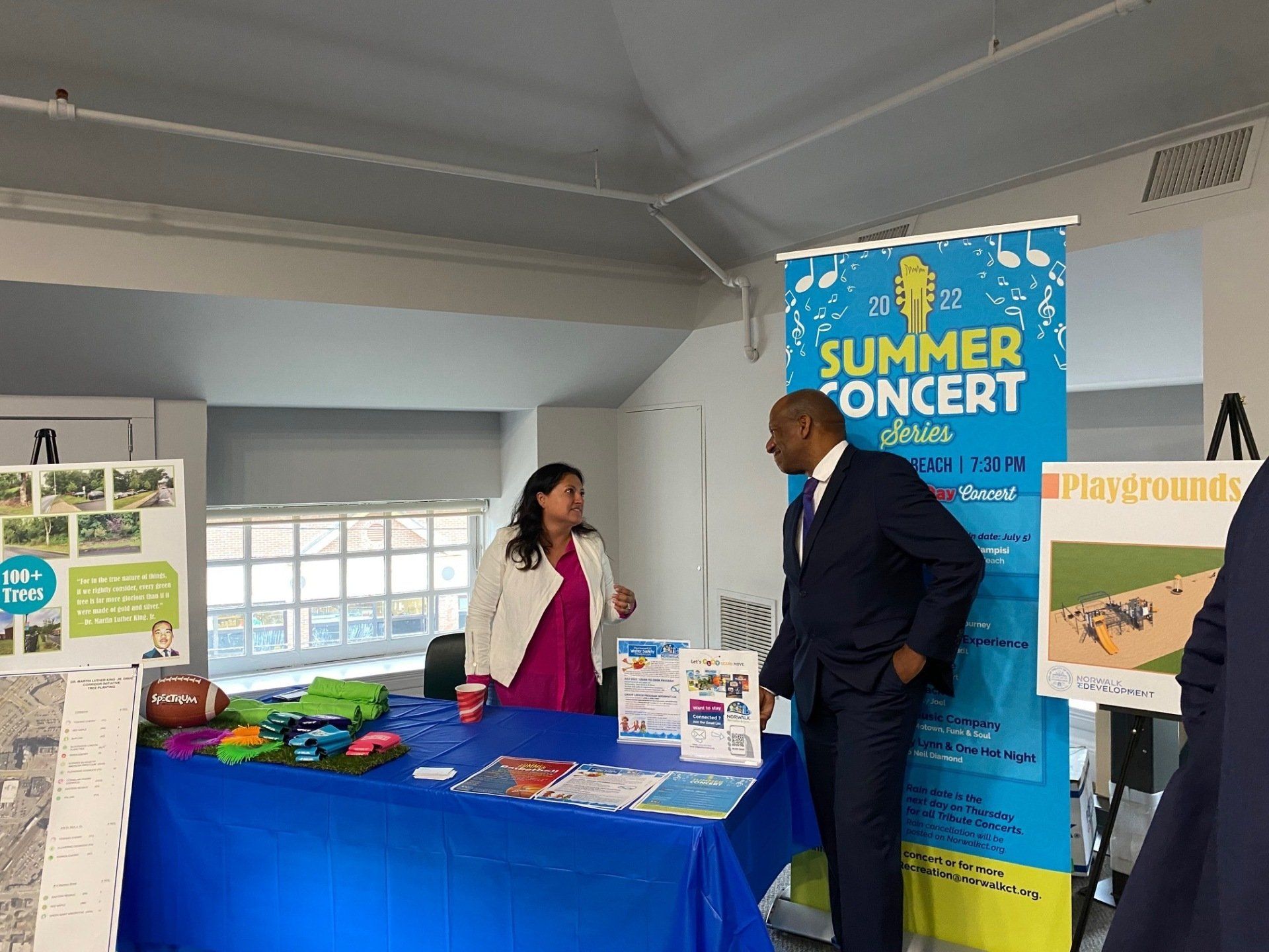 Two people stand at a table draped in blue, featuring event information and a Summer Concert Series sign.