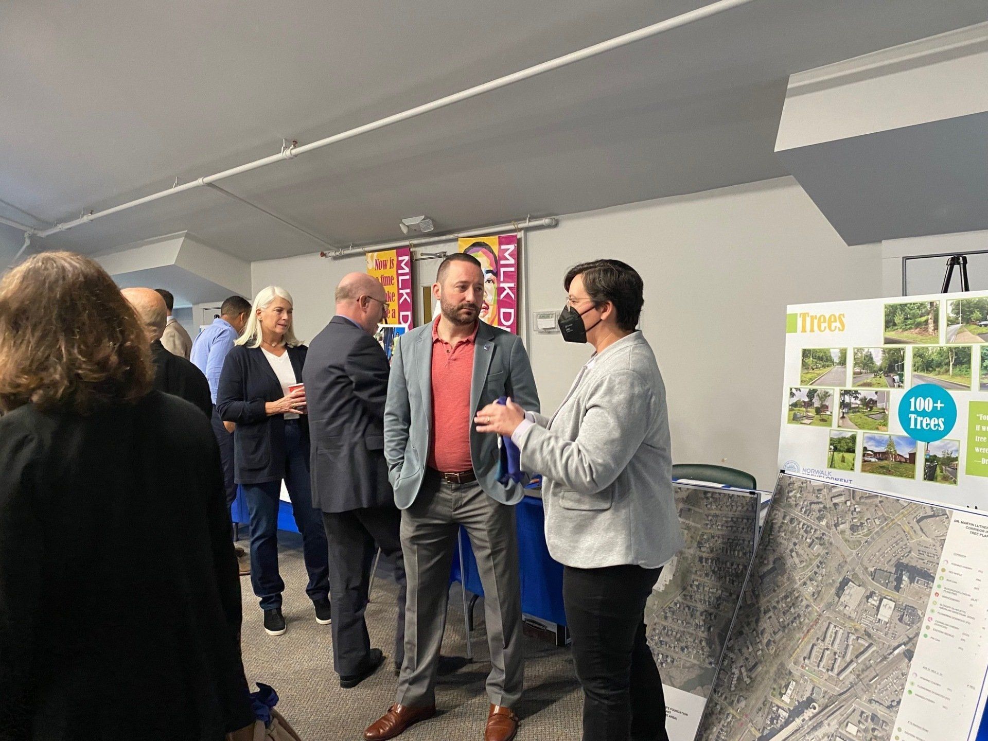 People stand and talk at an indoor community event with informational display boards featuring photos and a local map.