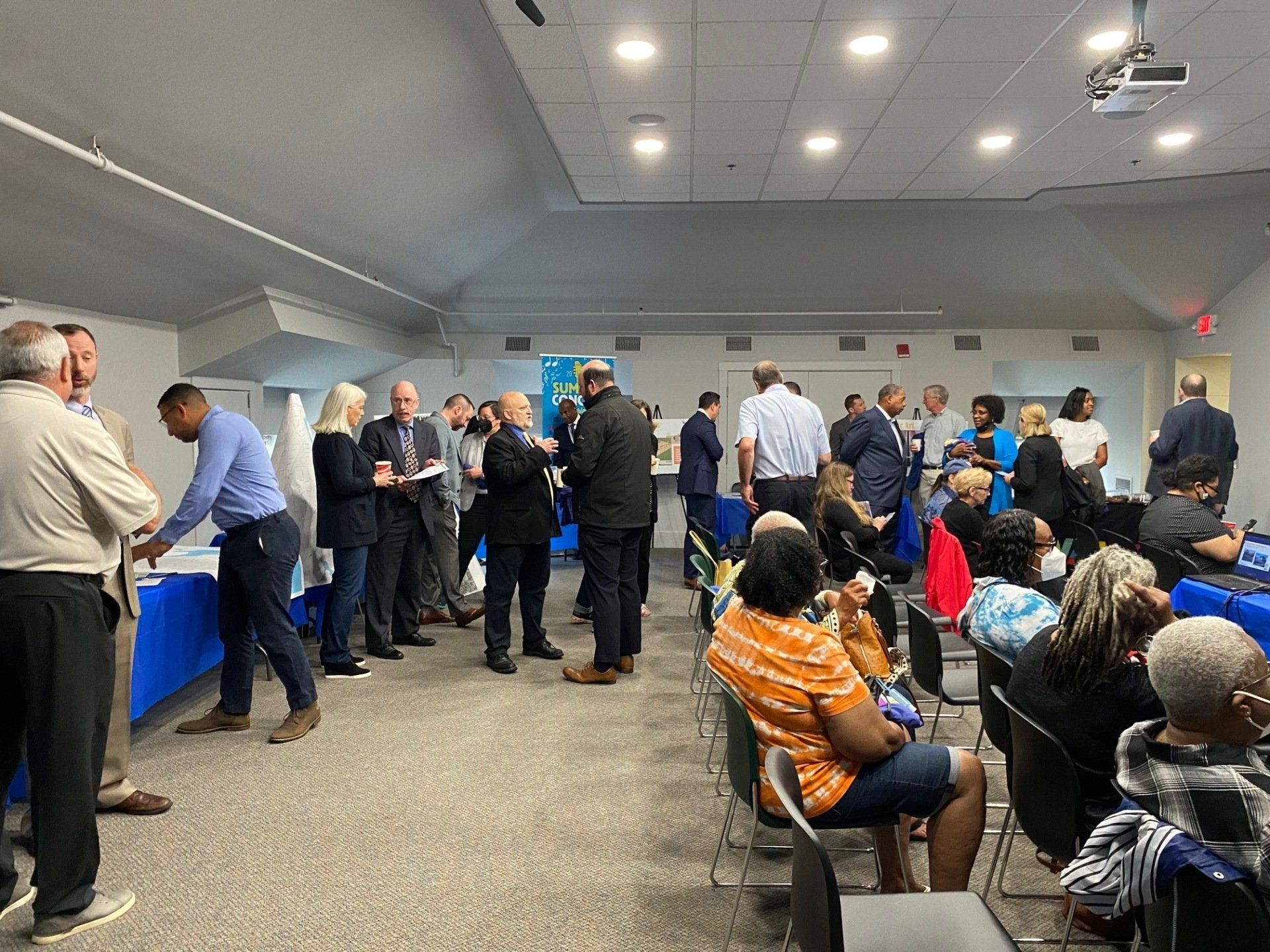People gather in a bright, spacious community room with blue-covered tables, likely attending a workshop or meeting.