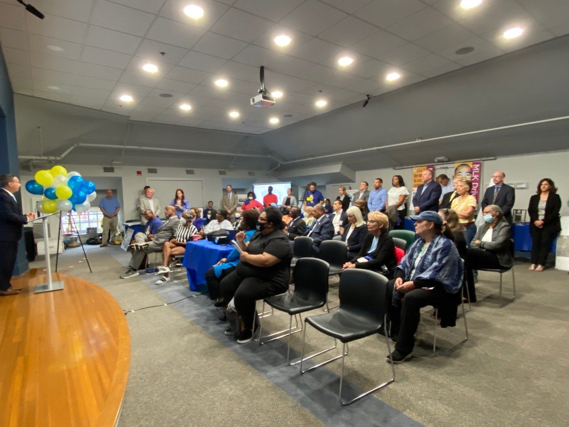 People attend a presentation in a room with a wooden stage, yellow and blue balloons, and rows of seating.