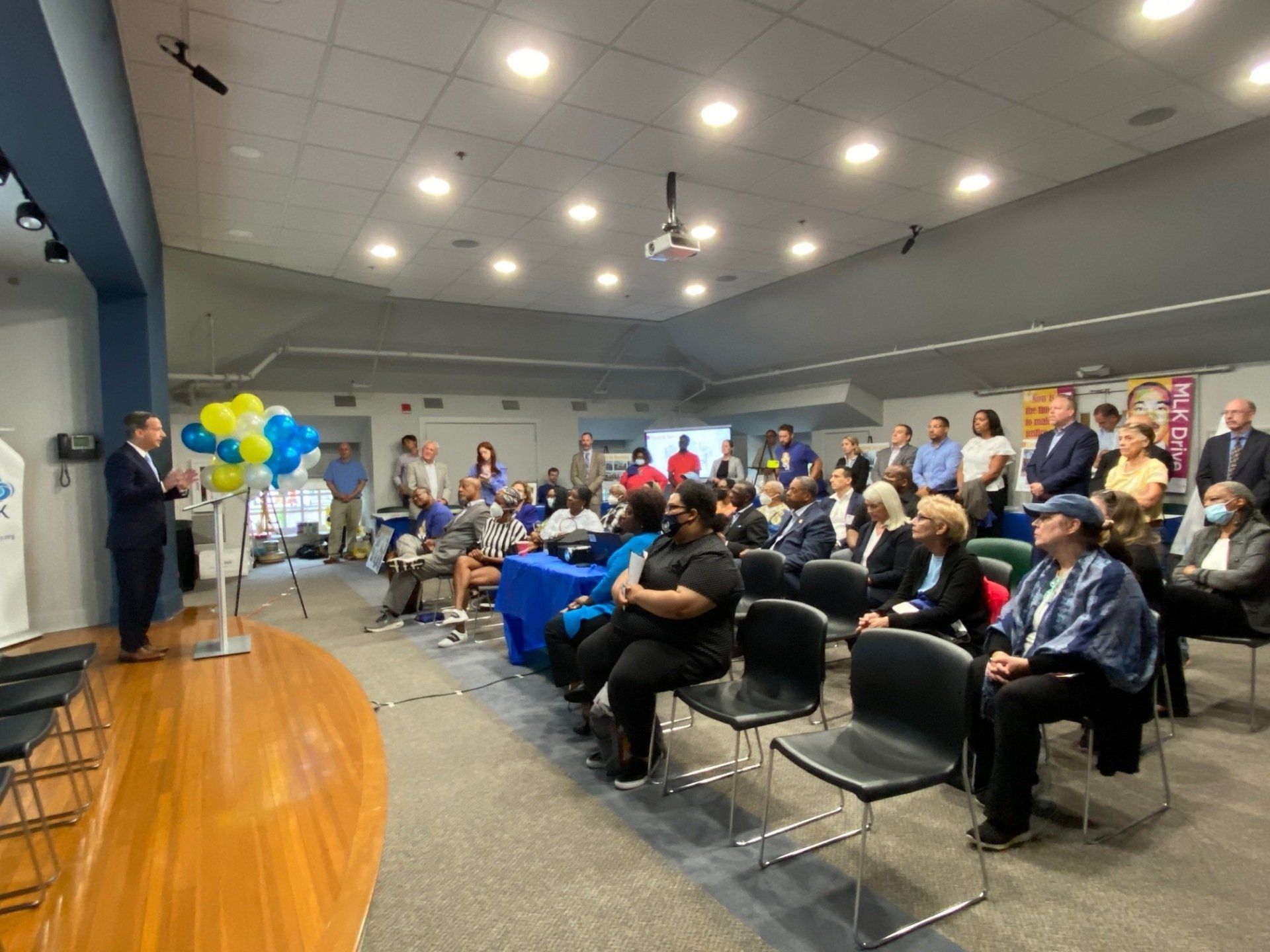 A speaker stands on a stage addressing a seated audience in a bright, indoor community or meeting room.