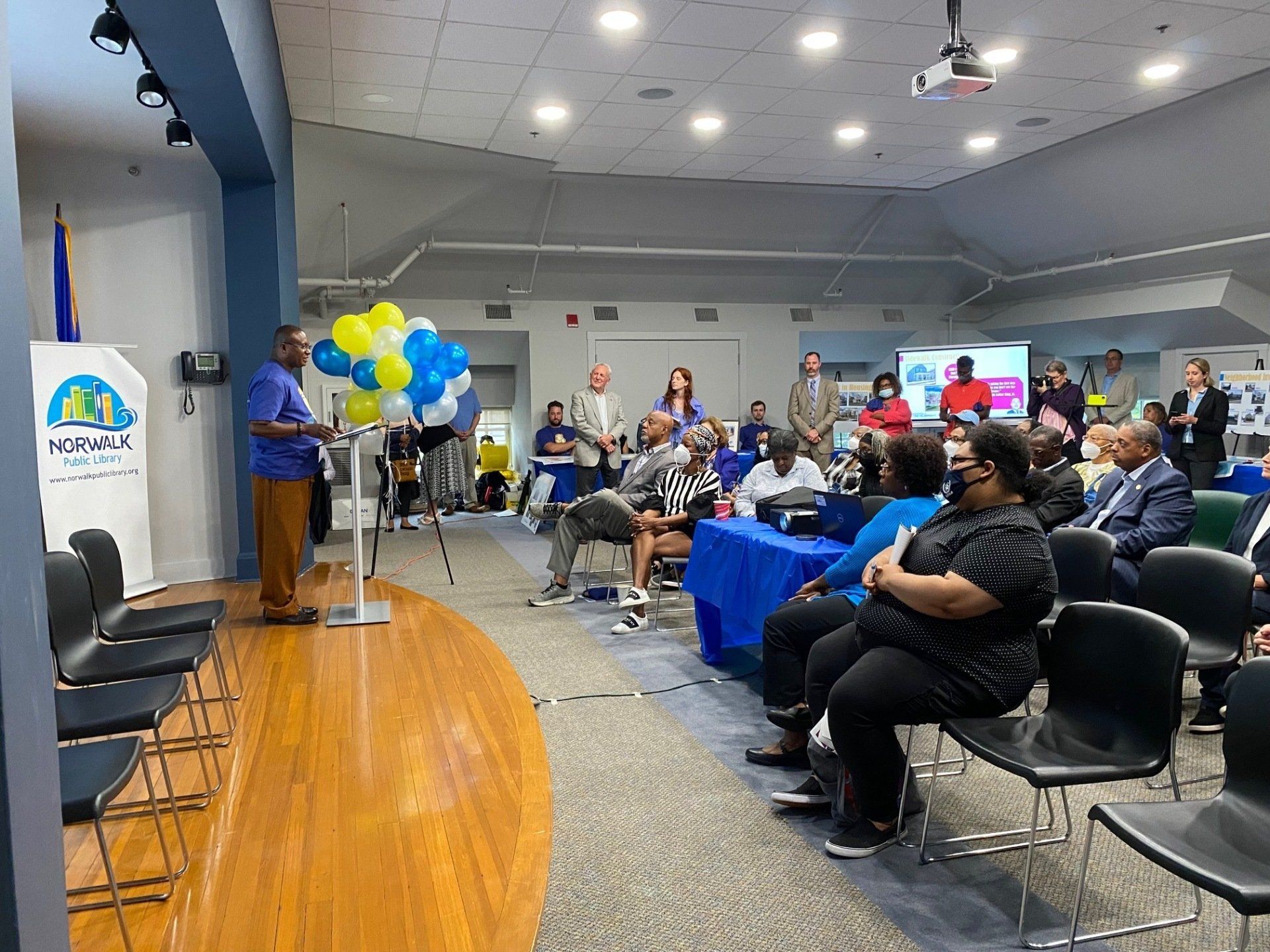 A speaker stands on a stage before an audience seated in a multipurpose room decorated with blue and yellow balloons.