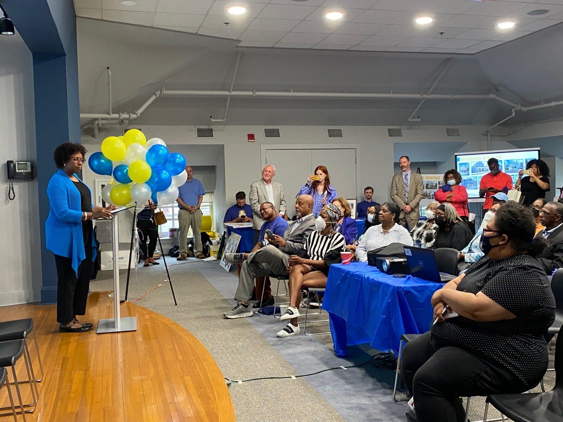 A person speaks from a podium to a diverse audience gathered in a community room with blue tablecloths and balloon decor.