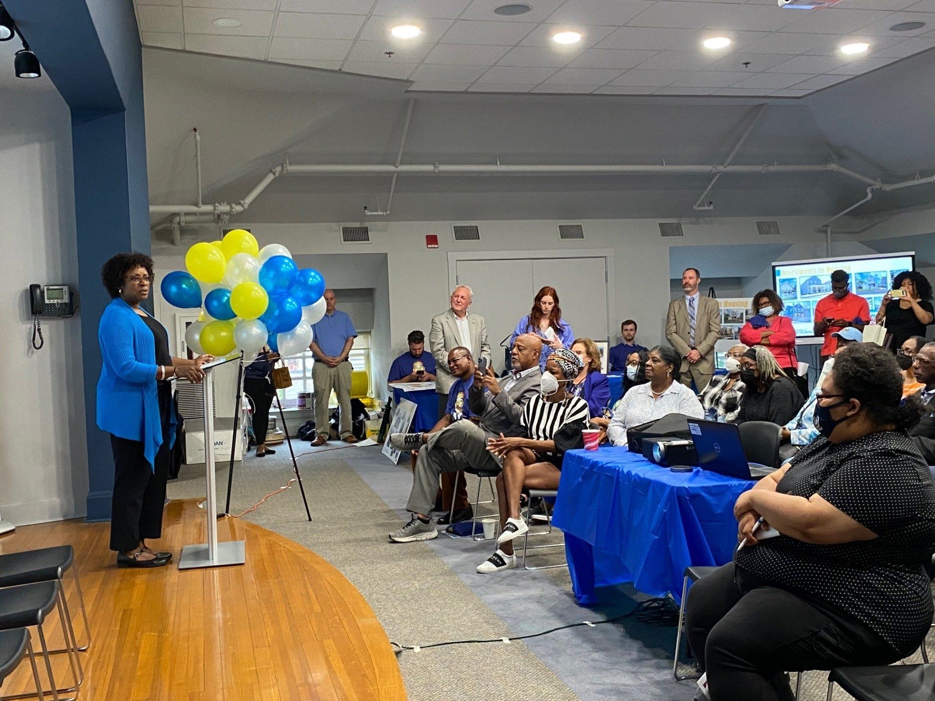 A person in a blue jacket speaks from a podium before an audience in a bright indoor room with yellow and blue balloons.