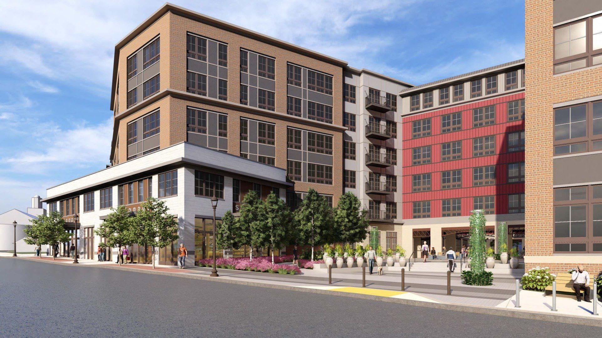 A modern multi-story apartment building with brick and red facades, featuring ground-level storefronts and a paved plaza.