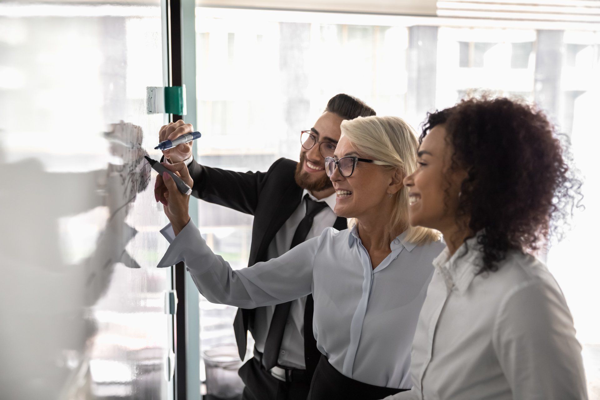 Three professionals collaborate at a glass wall, with one person writing on it while the others look on attentively.
