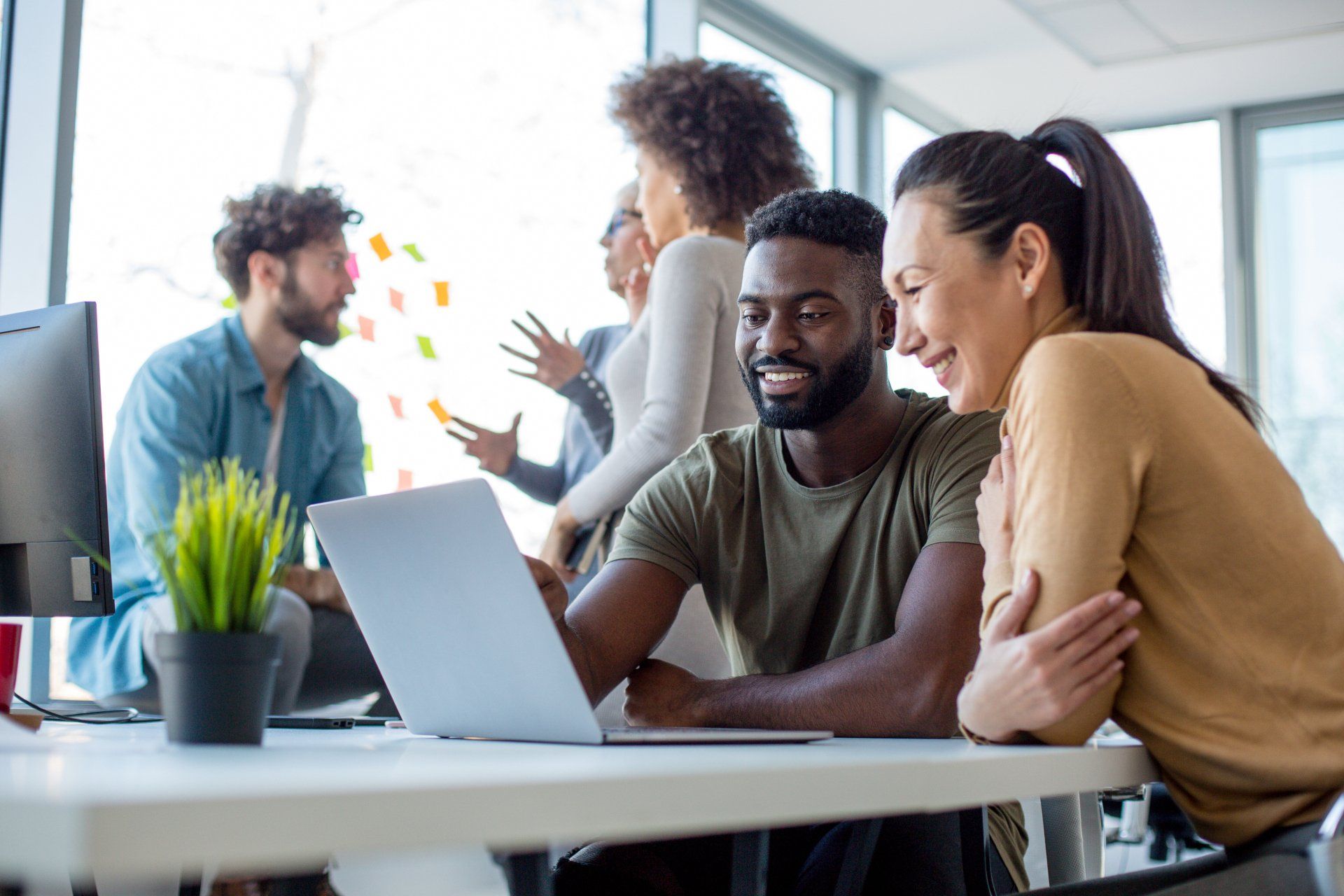 A diverse group of colleagues collaborating in a bright office; two people in the foreground smile while viewing a laptop.
