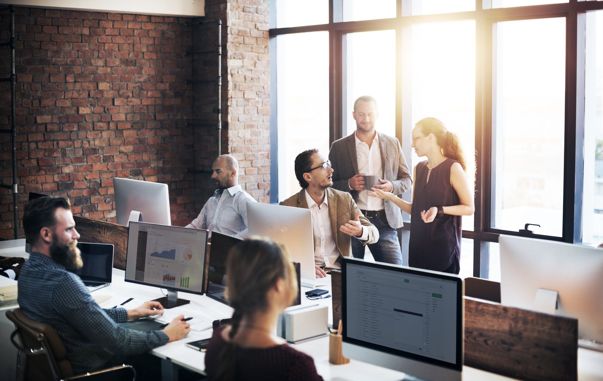 A team works in an open-plan office with a brick wall and large windows, some at desks and others standing in conversation.