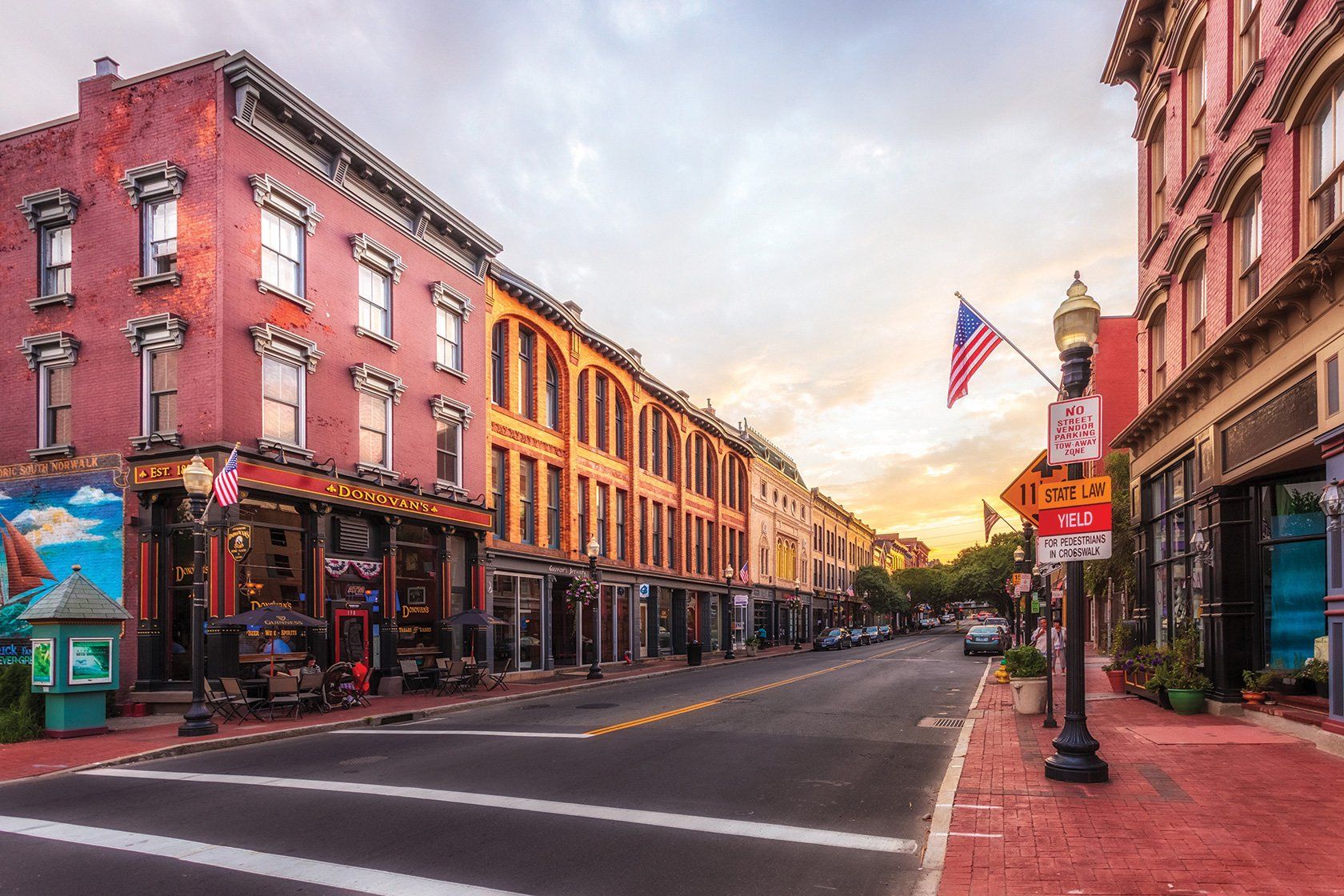 A scenic city street with red and tan historic buildings at sunset, featuring an American flag and a street lamp.