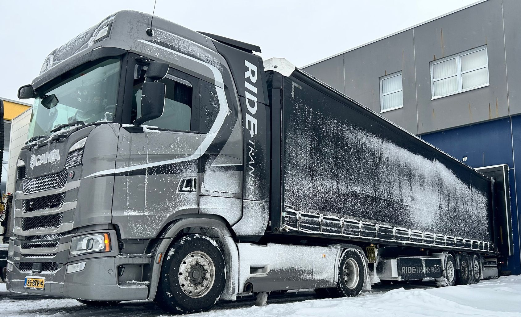 Grijze, met sneeuw bedekte vrachtwagen geparkeerd voor een gebouw tijdens een sneeuwbui.