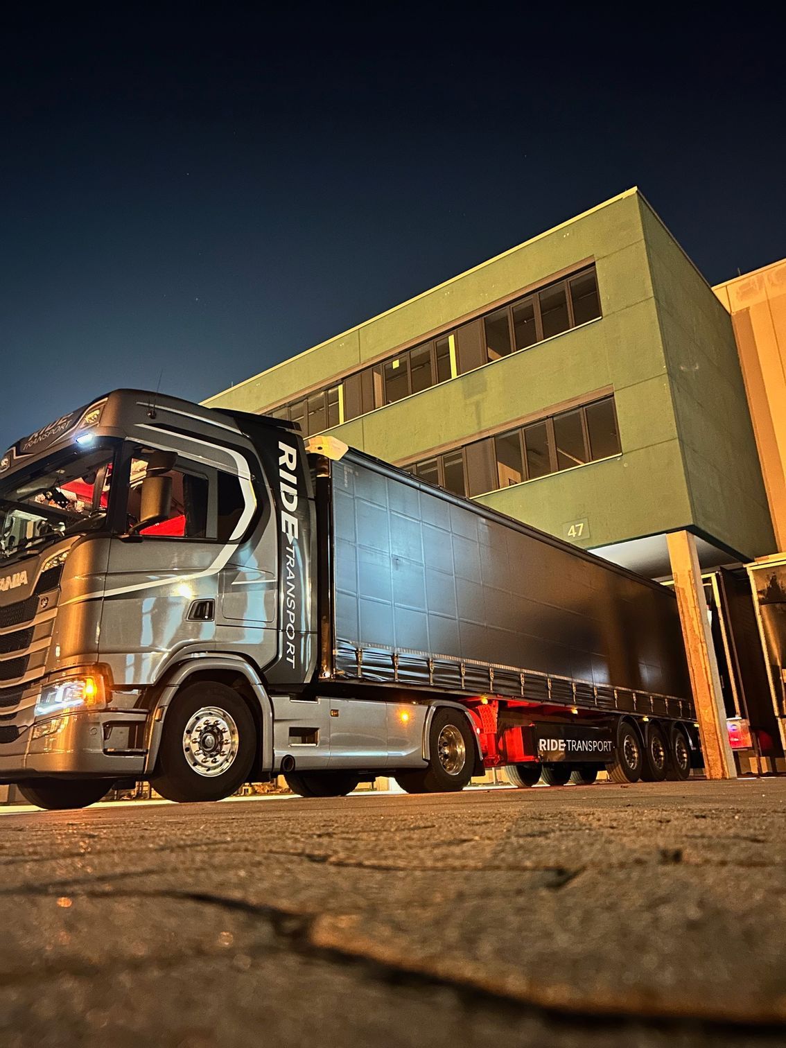 Een zilverkleurige vrachtwagen staat 's nachts geparkeerd naast een geel gebouw. De oplegger is verlicht.