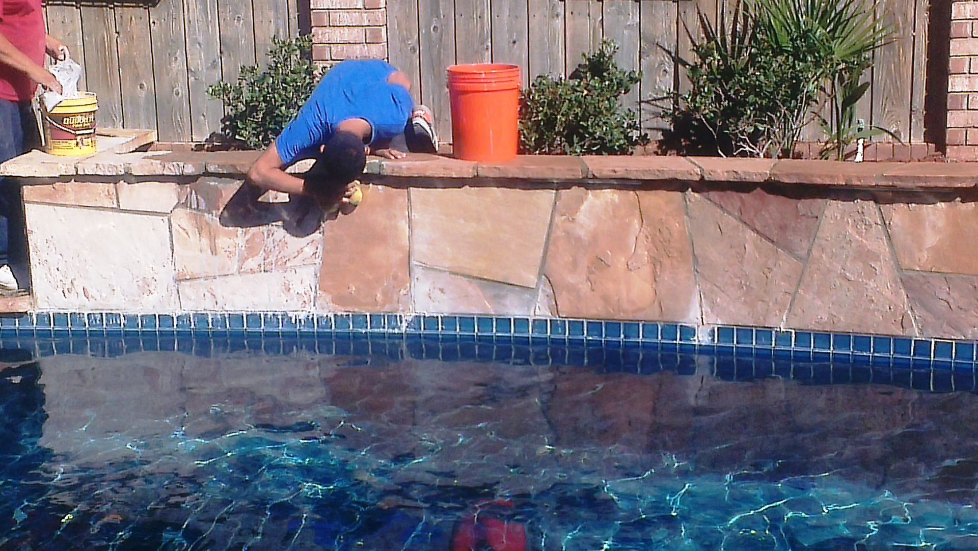 Man working on pool coping, leaning over pool edge. Orange bucket, blue tile, water, brick wall.